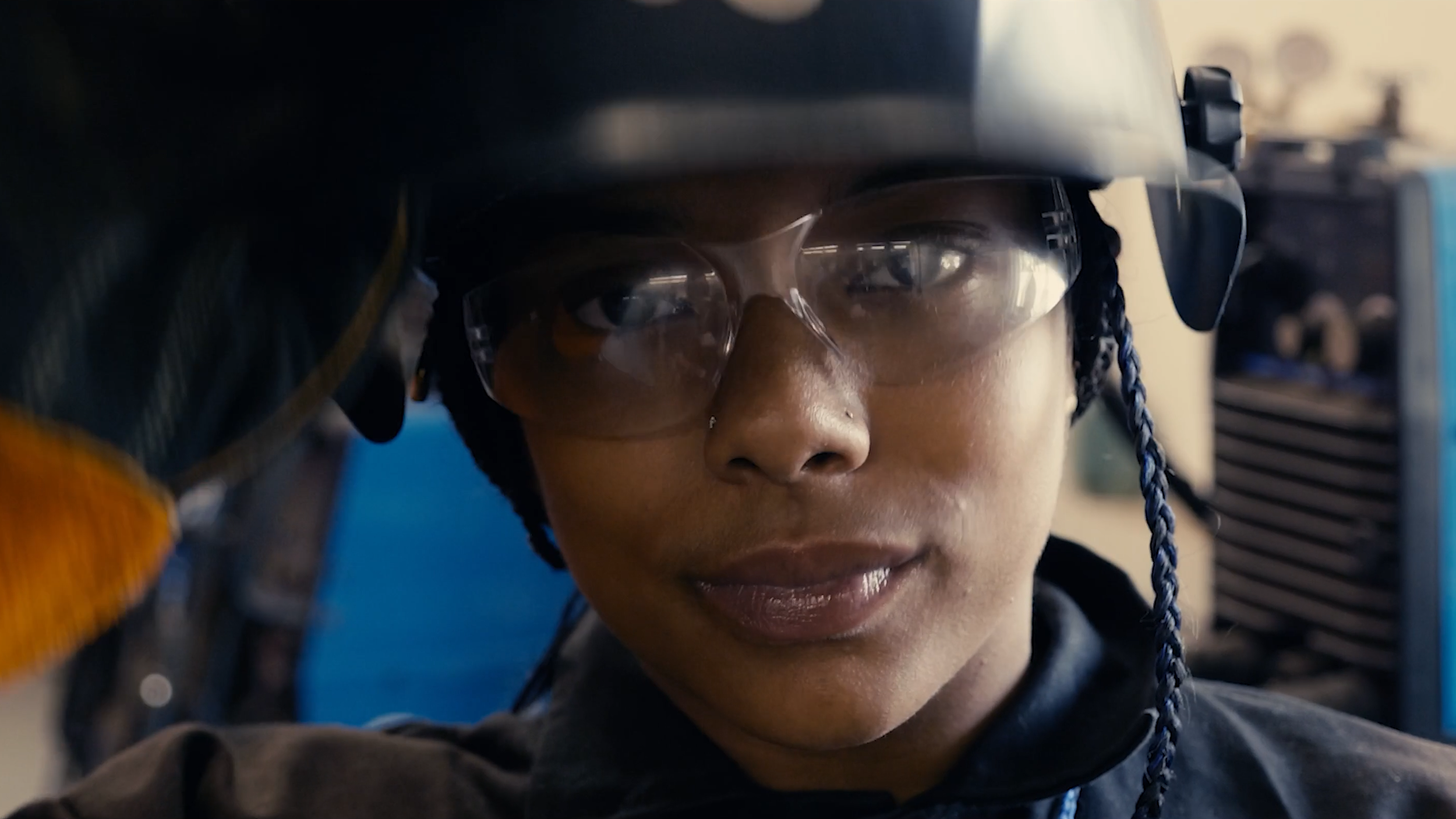 Close-up of a person with glasses and braided hair, wearing a helmet and protective gear, focusing on their face.