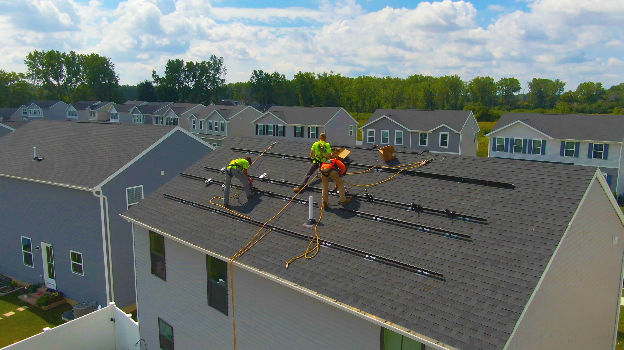 Workers installing solar panels on the roof of a multi-story house in a suburban neighborhood with other houses and trees in the background.