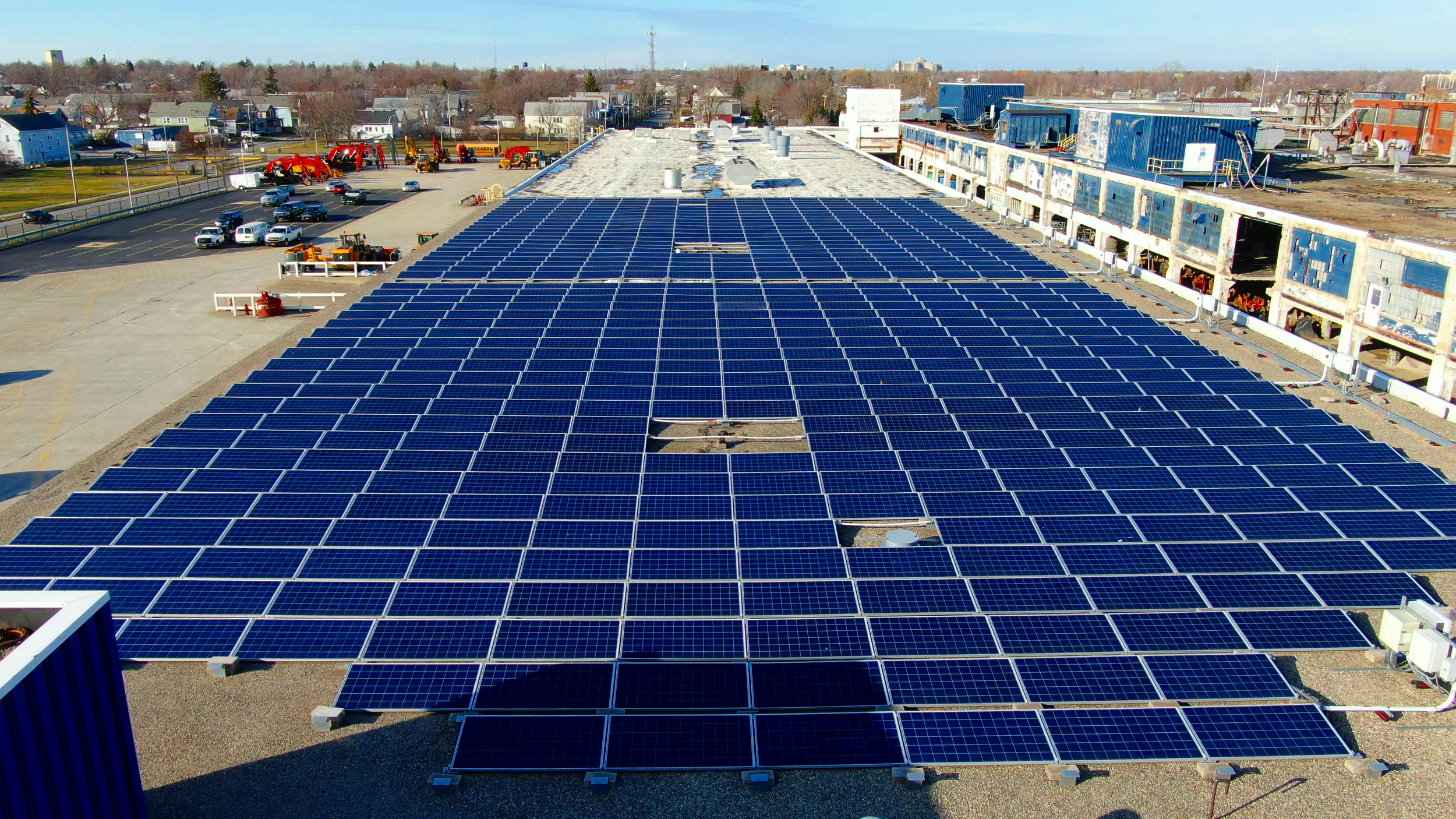 An aerial view of a large rooftop covered with numerous solar panels, with a parking lot and residential buildings in the background.