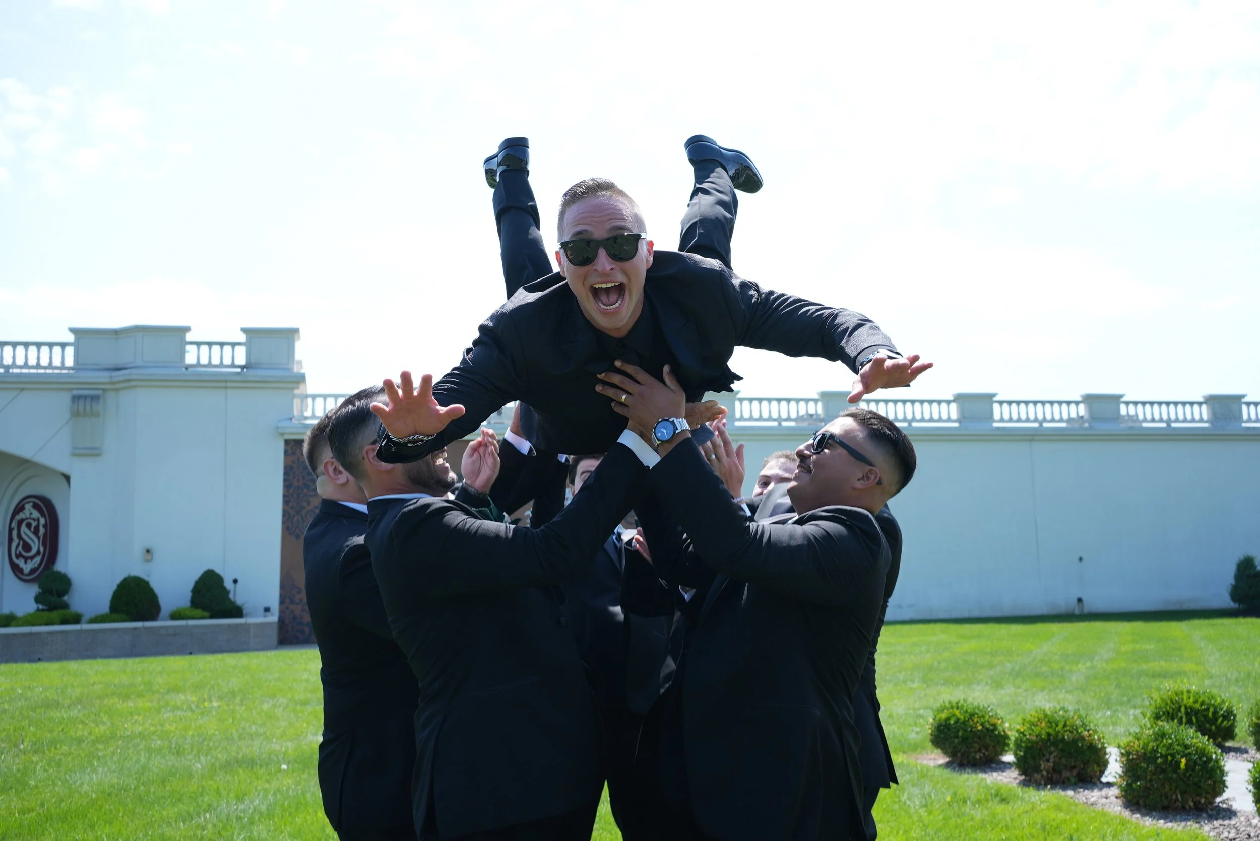 A group of men in suits holding up a man in a black suit and sunglasses, who is smiling and has his arms outstretched in celebration outside on a green lawn with a white building in the background.