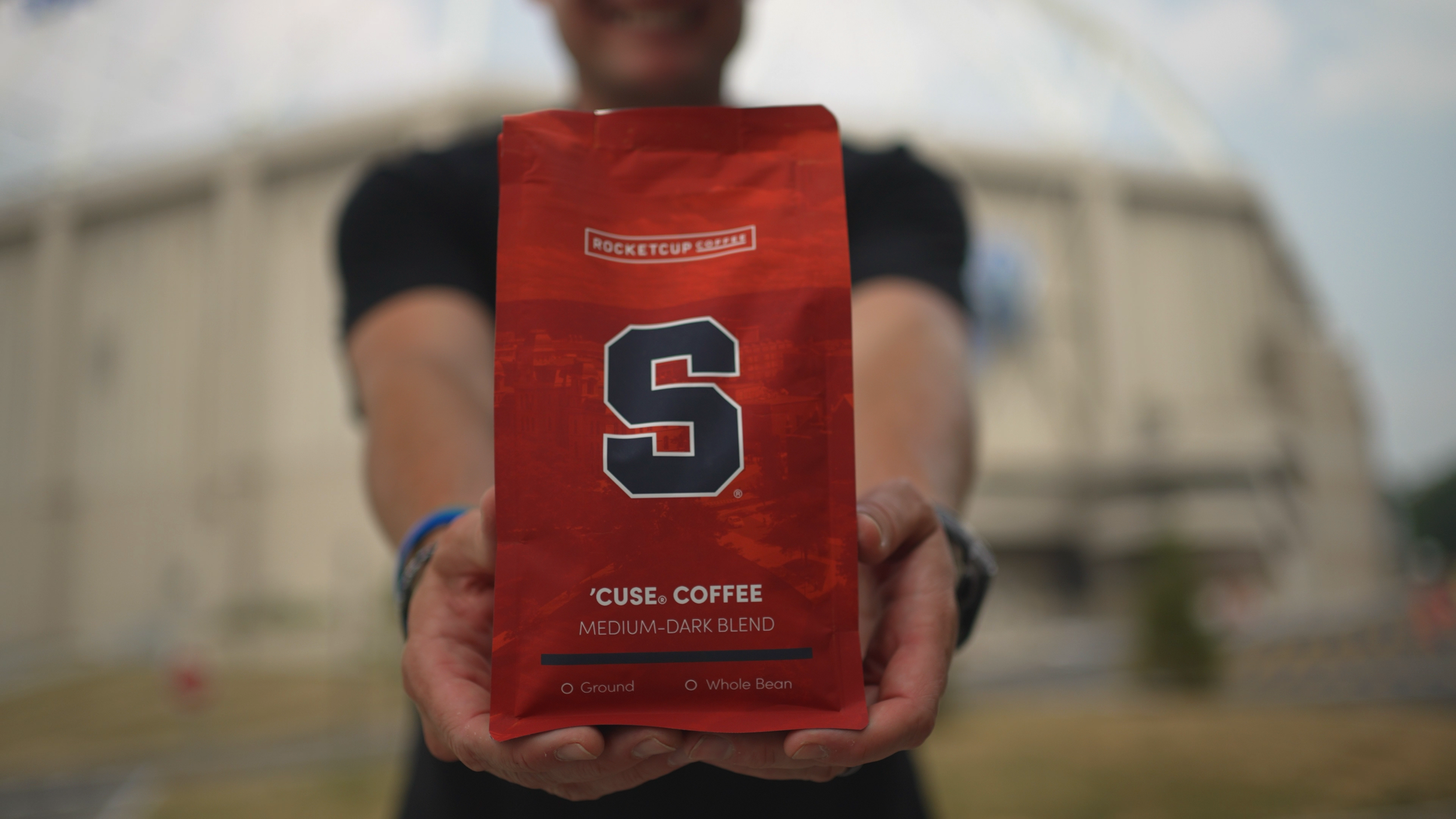 Person holding a red bag of Rocket Fuel Coffee branded 'S' logo with text 'Cuse Coffee' and 'Medium-Dark Blend' in front of a blurred background.