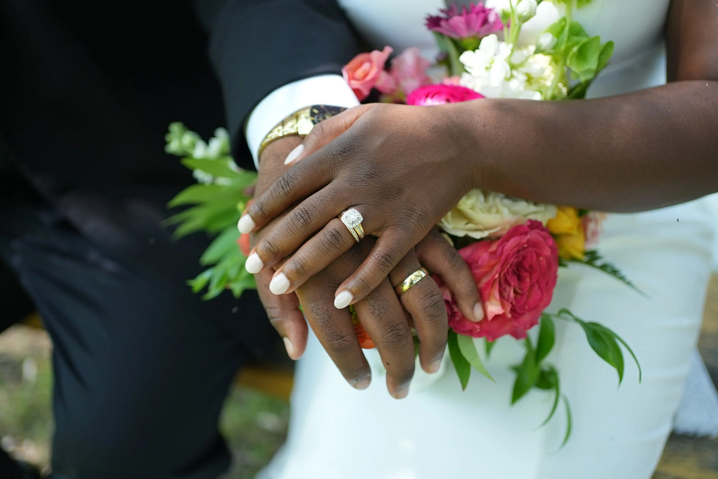 Close-up of a couple holding hands, both wearing wedding rings, with a bouquet of pink and white flowers in the background.