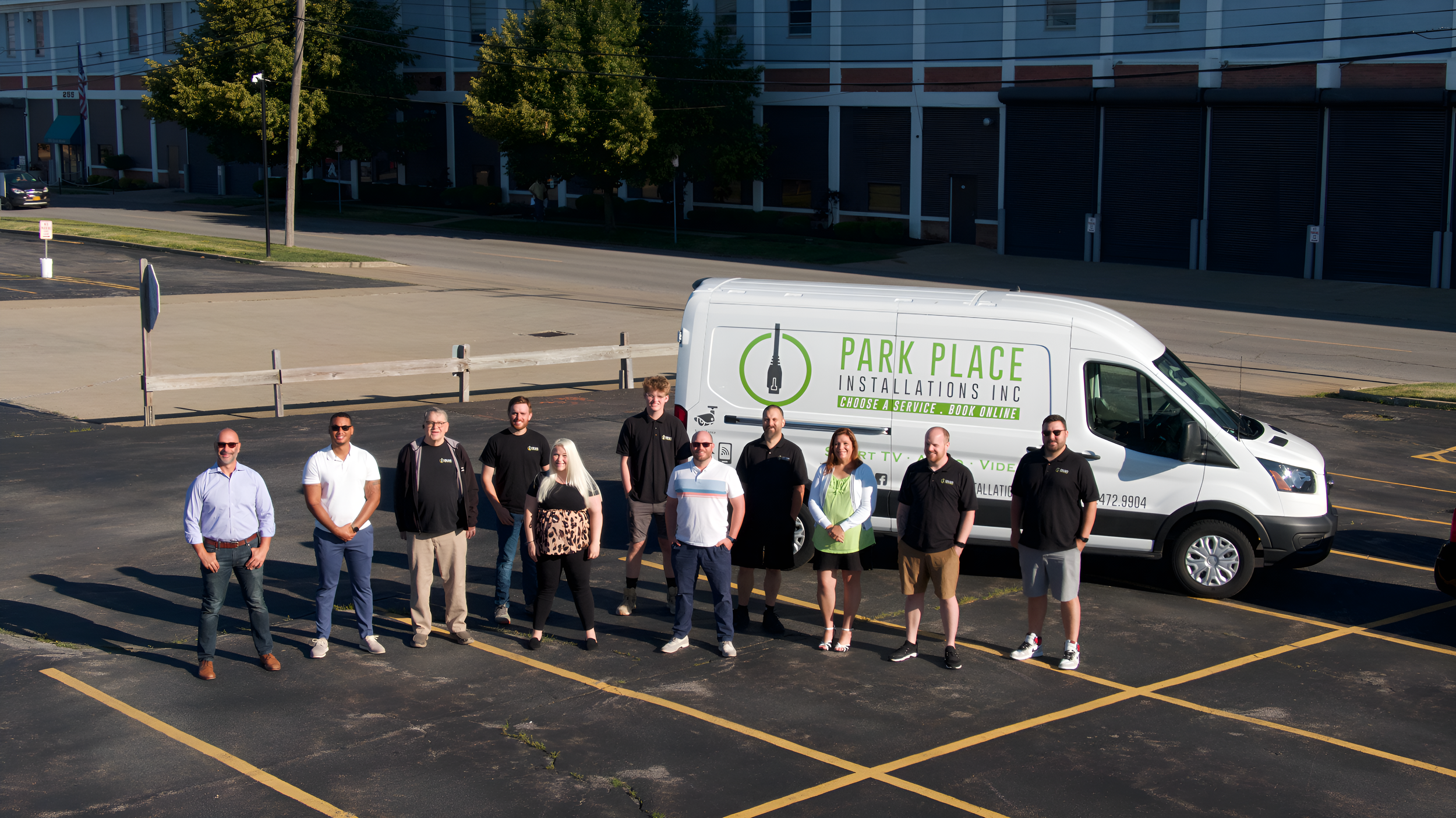 Group of eleven people standing in parking lot, with company van labeled 'Park Place Installations Inc' behind them, in an urban setting with trees and buildings in the background.