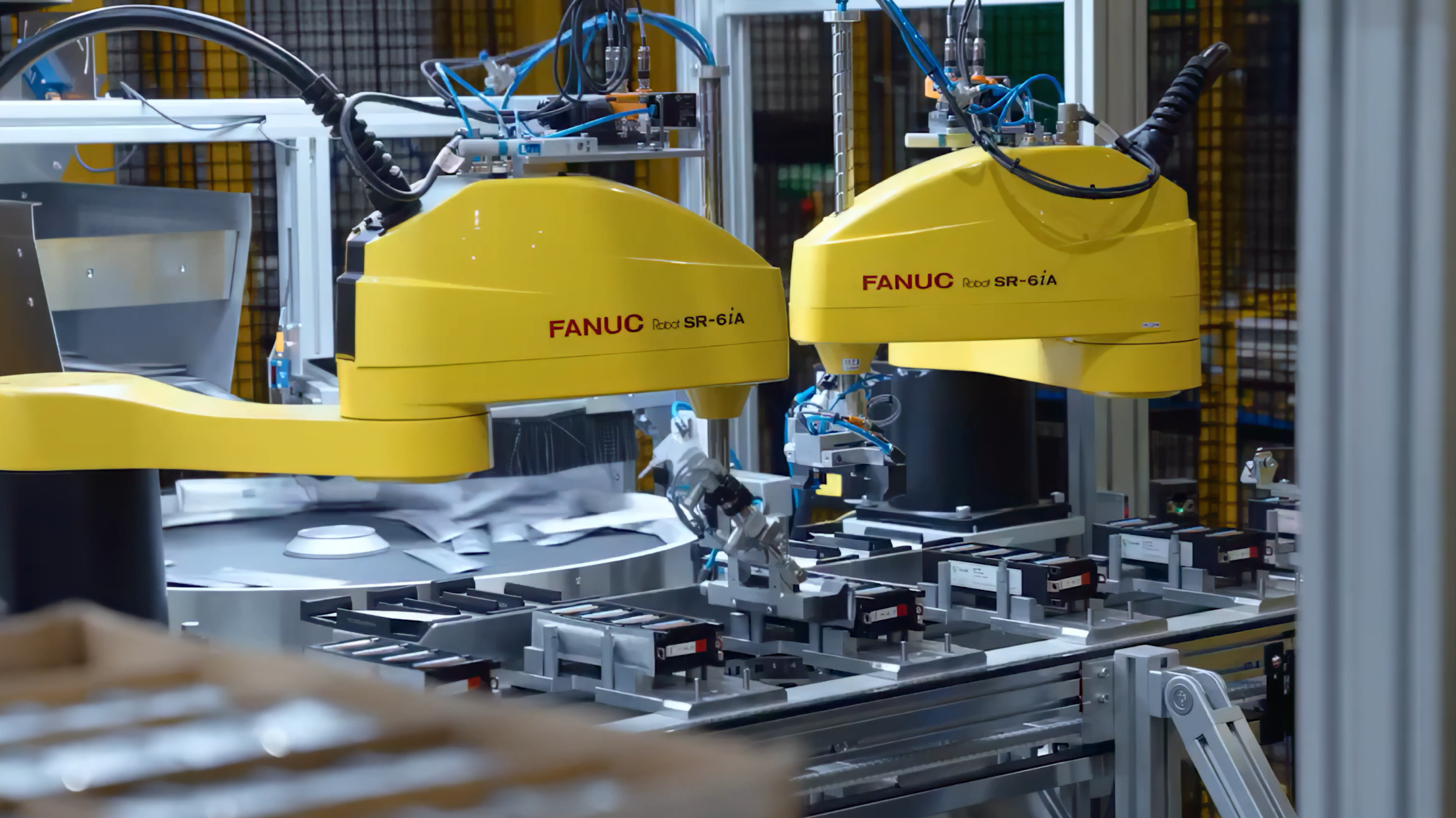 Two yellow FANUC robotic arms operating on a manufacturing assembly line with electronic components and machinery in the background.