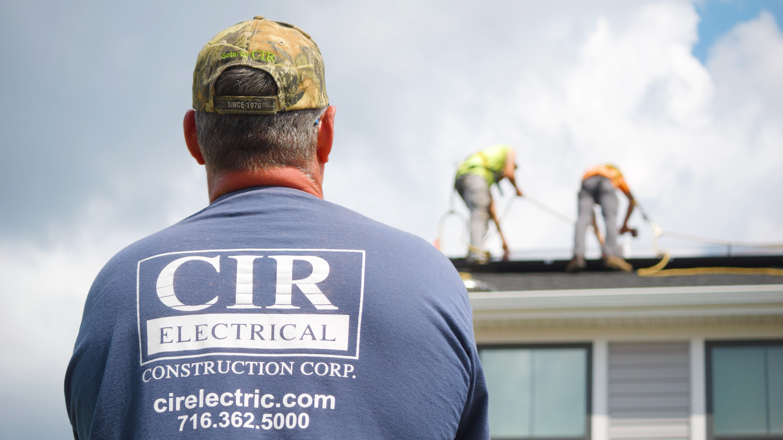 A man wearing a blue shirt and camouflage cap watches two workers on a roof, with the company's logo and contact information on his shirt.