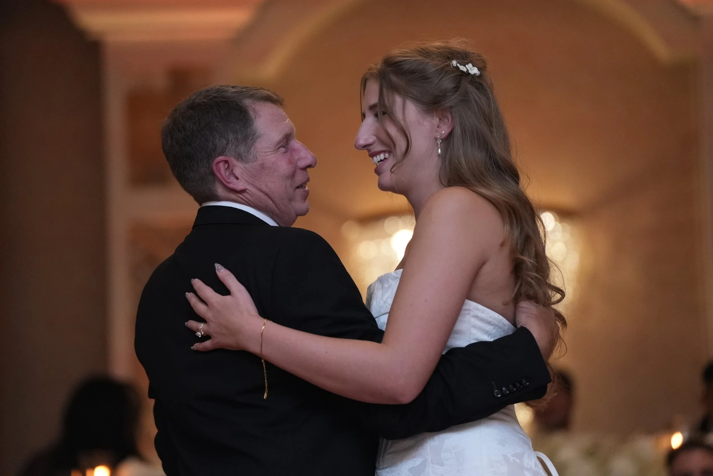A man in a black tuxedo and a woman in a white dress dancing closely at a wedding reception, smiling and looking into each other's eyes.