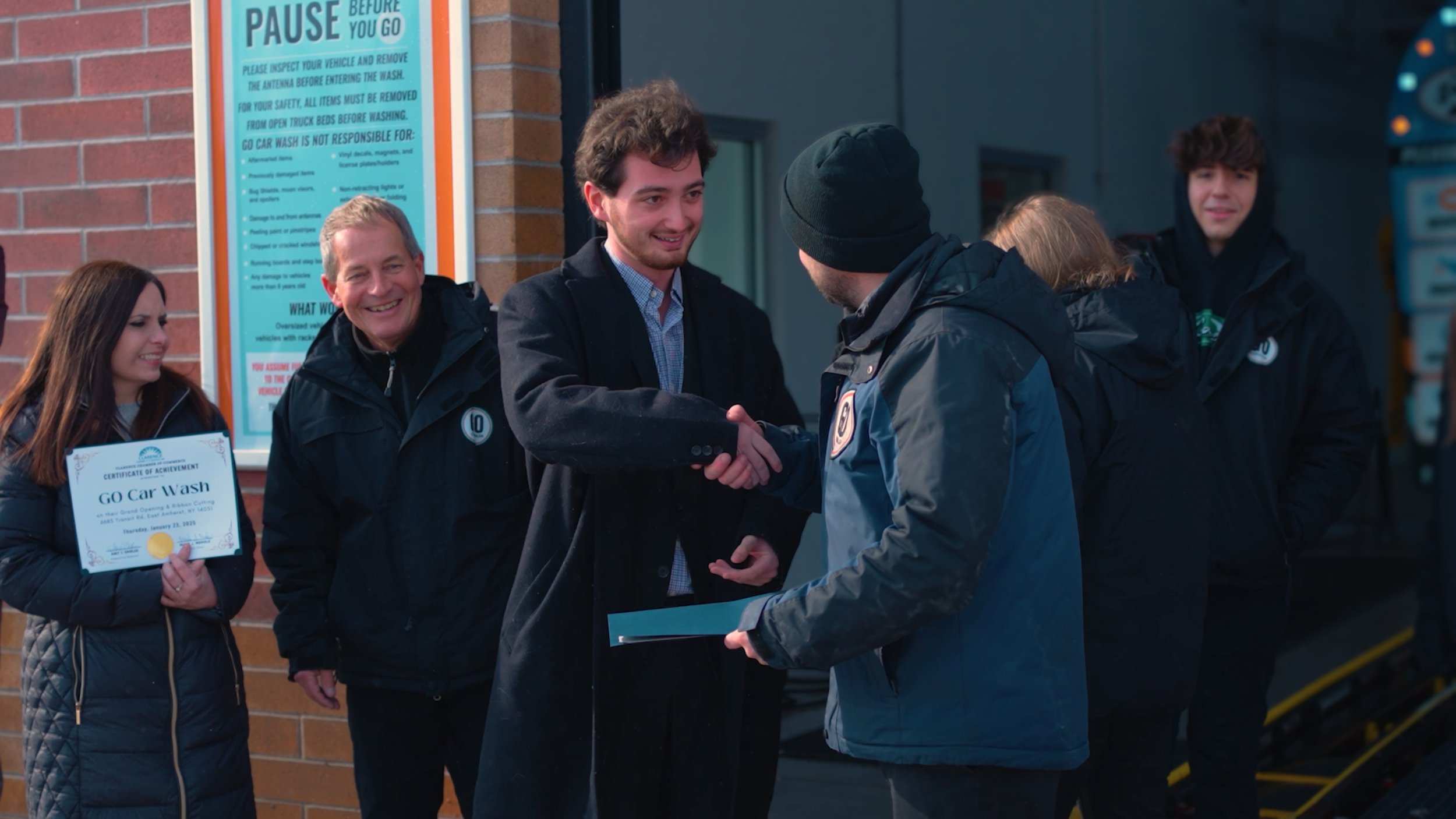 Group of people at a grand opening event for a car wash, with a man shaking hands with another person and others smiling, holding a certificate of achievement for the car wash.