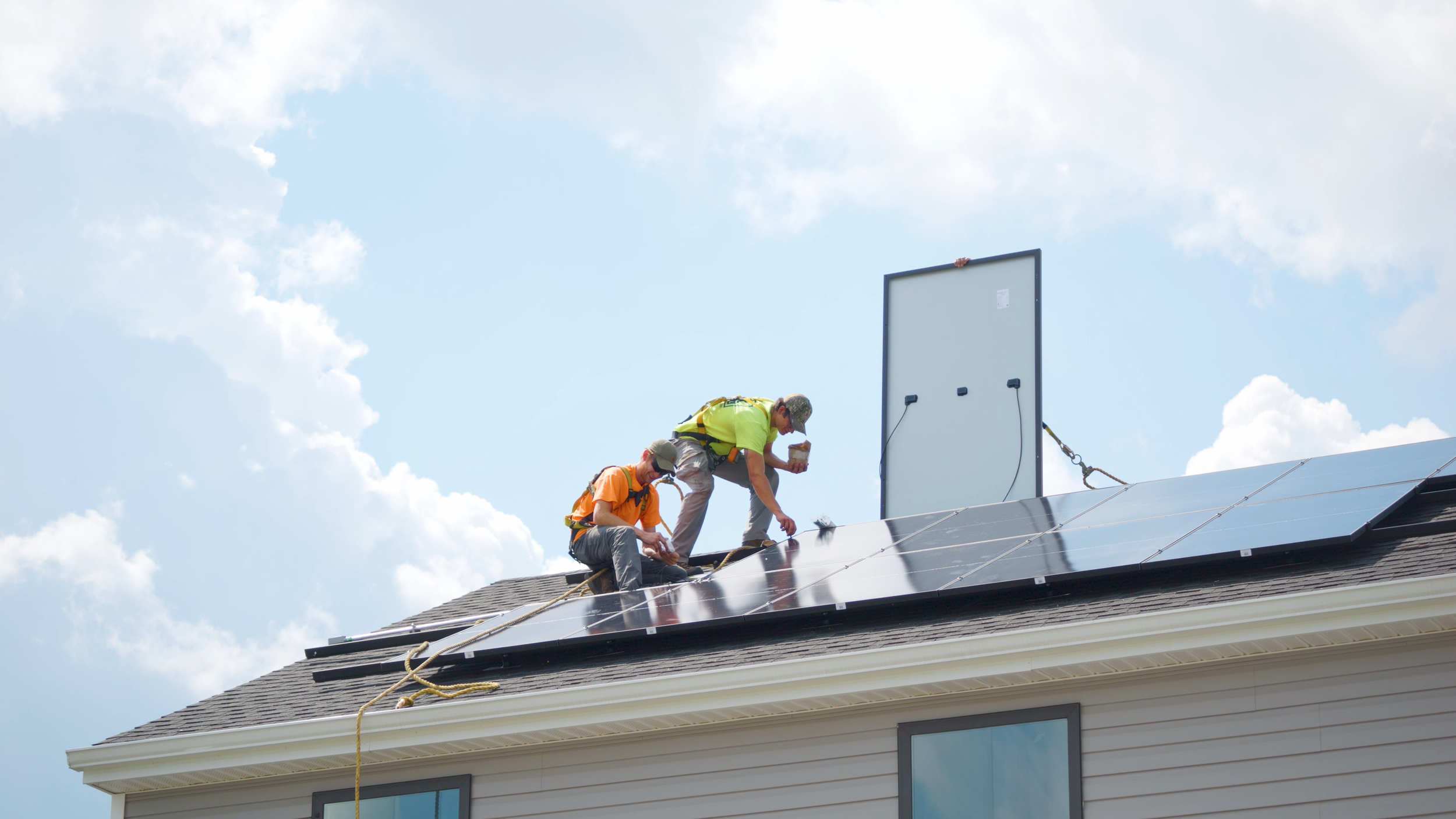 Two workers installing solar panels on a residential roof during the daytime, with a clear sky and clouds in the background.