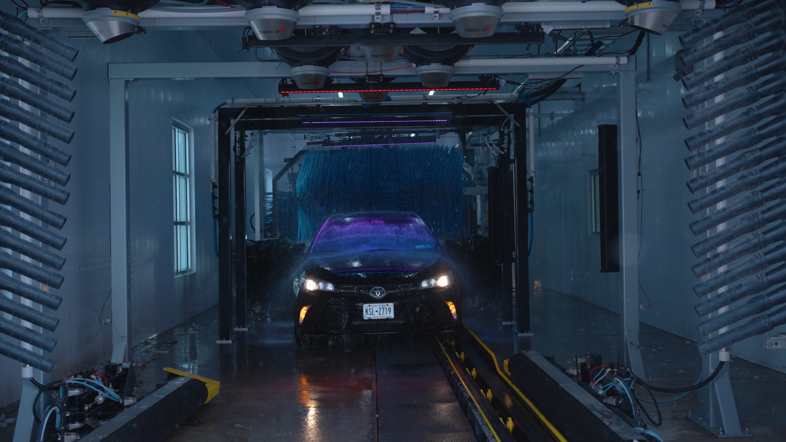 Automated car wash with a black vehicle being rinsed with water under a robotic washing system inside a white-walled room with windows.