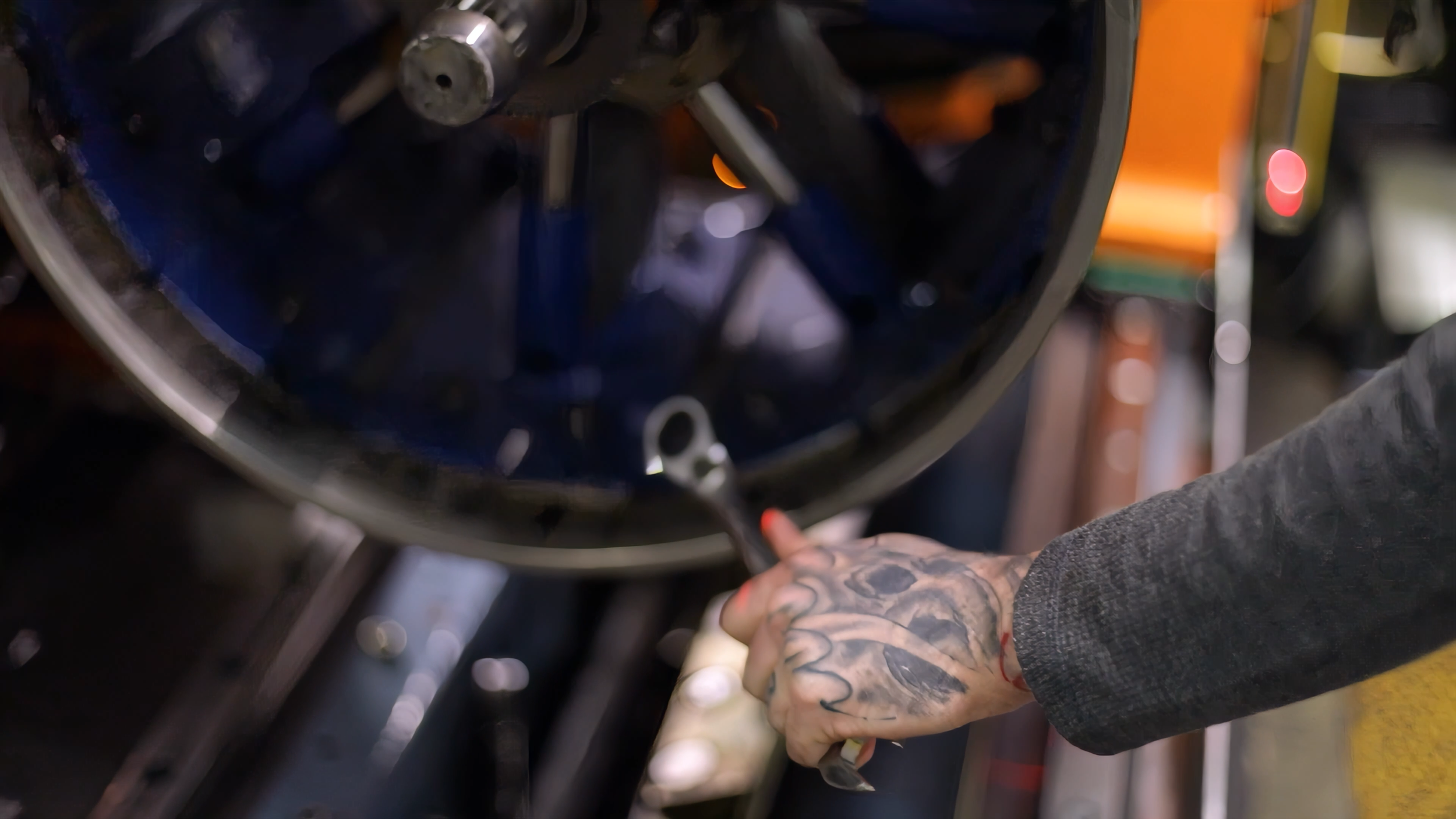 Close-up of a person with tattooed hands working on a mechanical component, possibly in a workshop or garage.