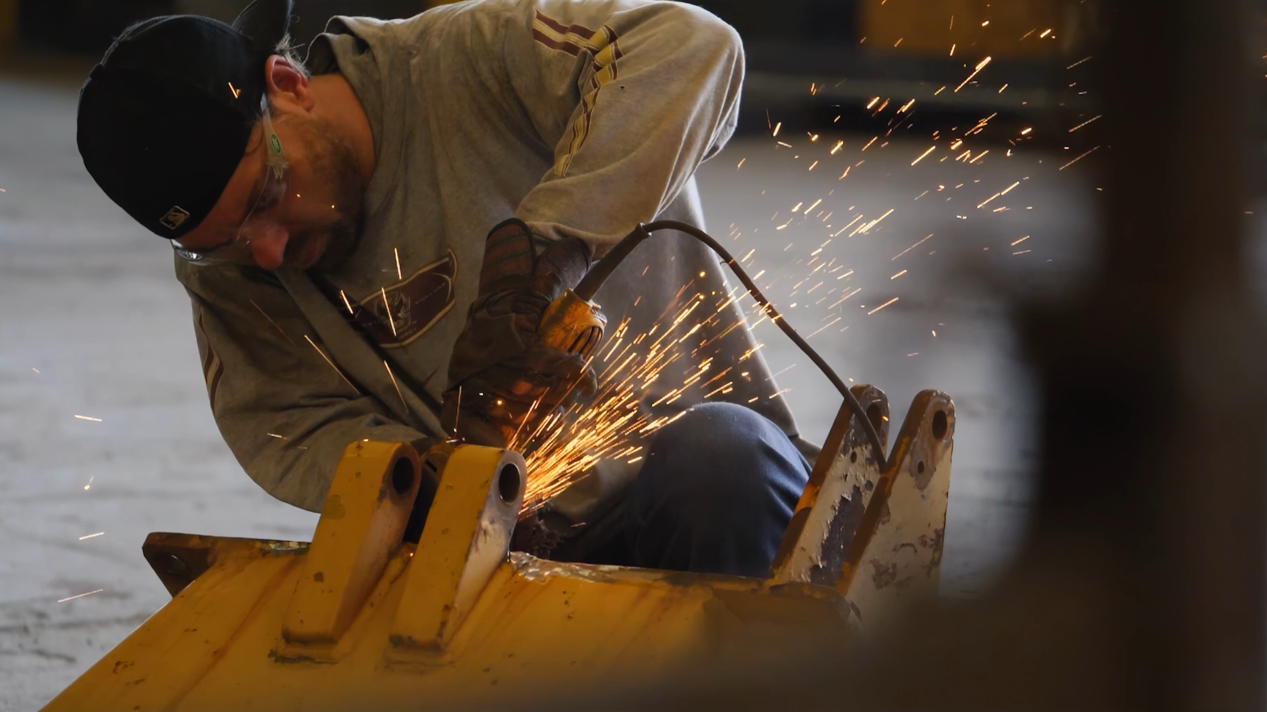 A man welding metal part, sparks flying around him, sitting on a yellow metal structure in an industrial setting.