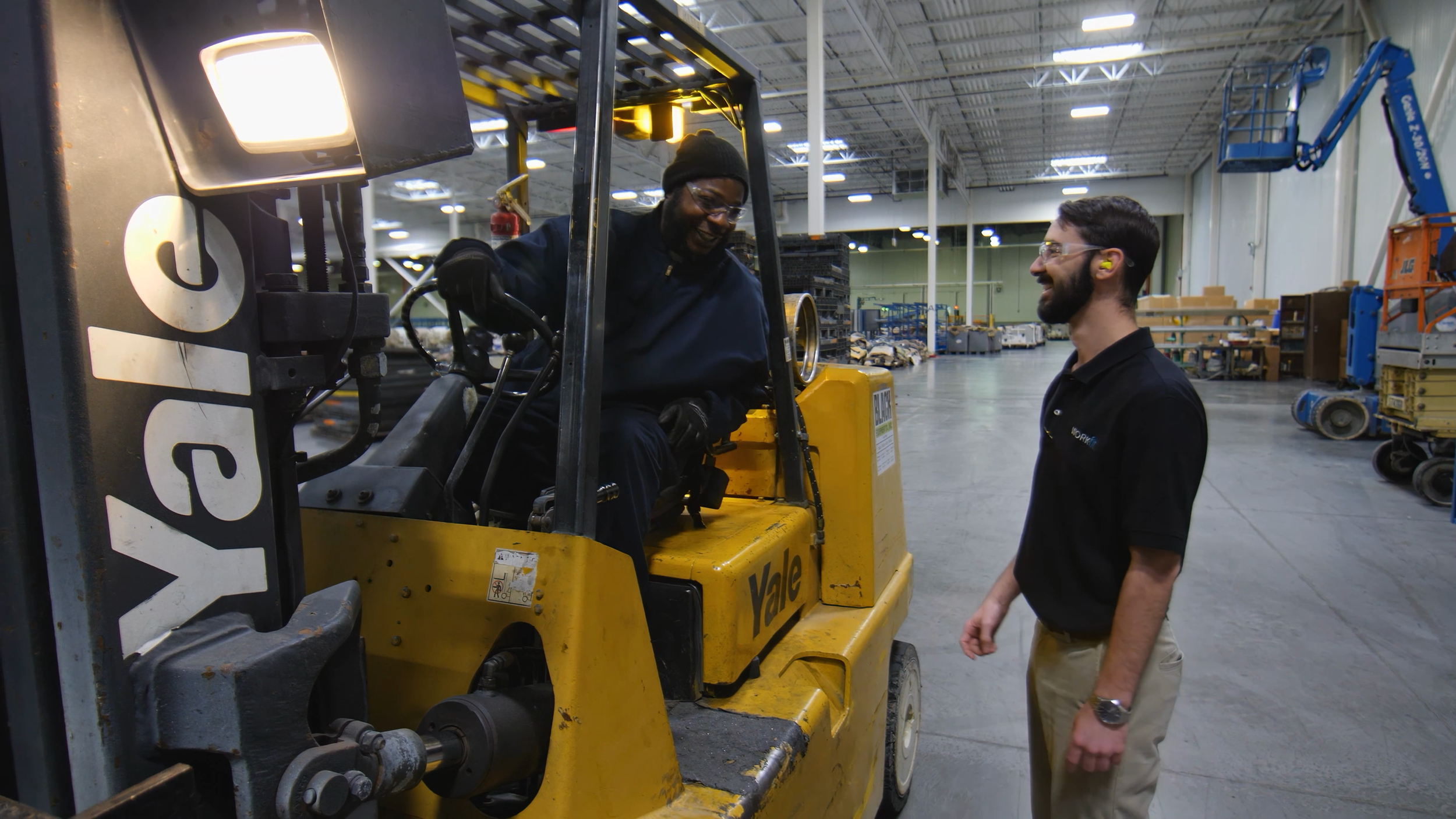 Two men, one sitting on a yellow forklift and the other standing next to it, are having a conversation inside a warehouse with various equipment and shelves in the background.