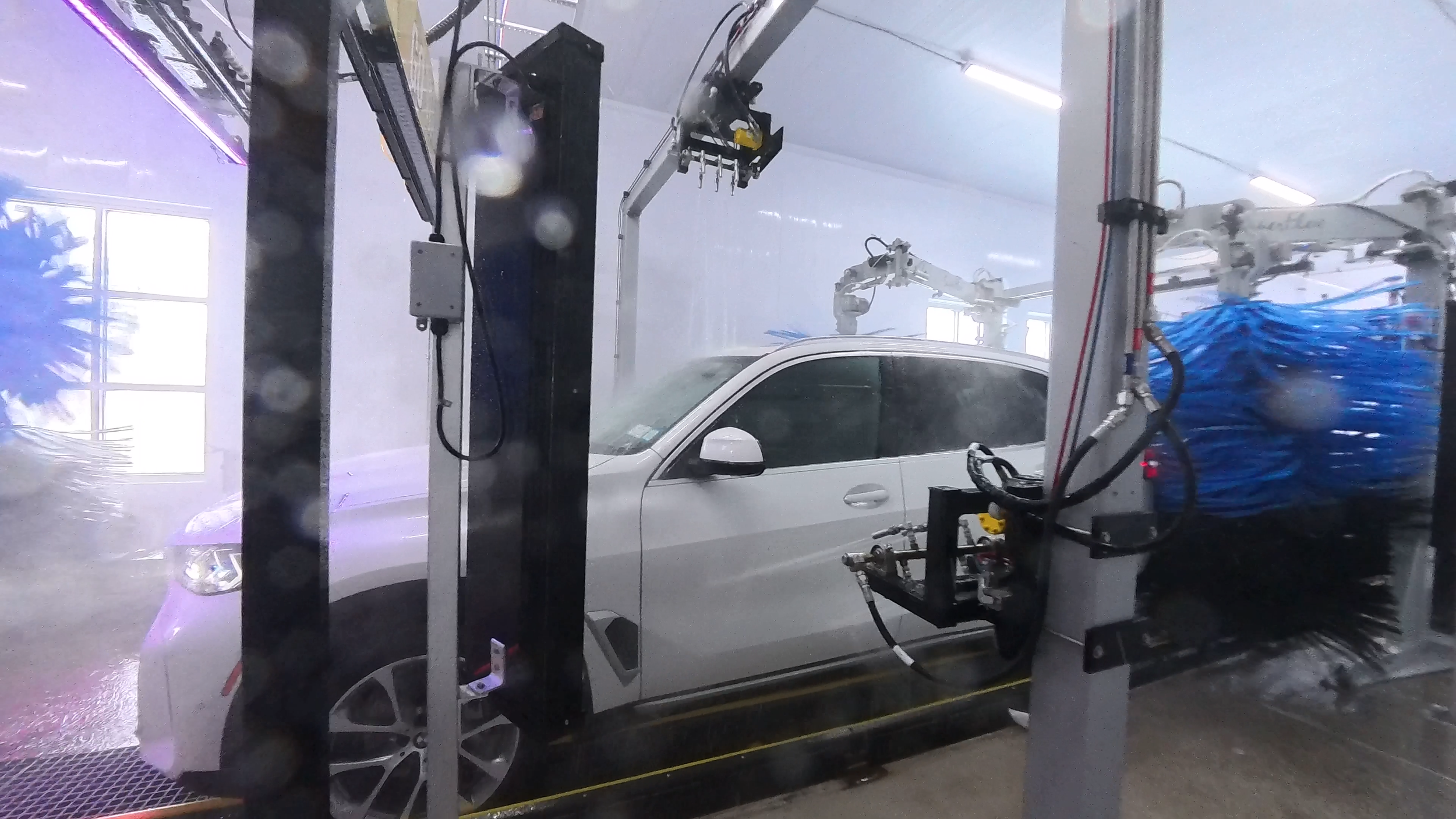 An indoor car wash facility with a white vehicle being cleaned by automated robotic arms, with water and soap visible.