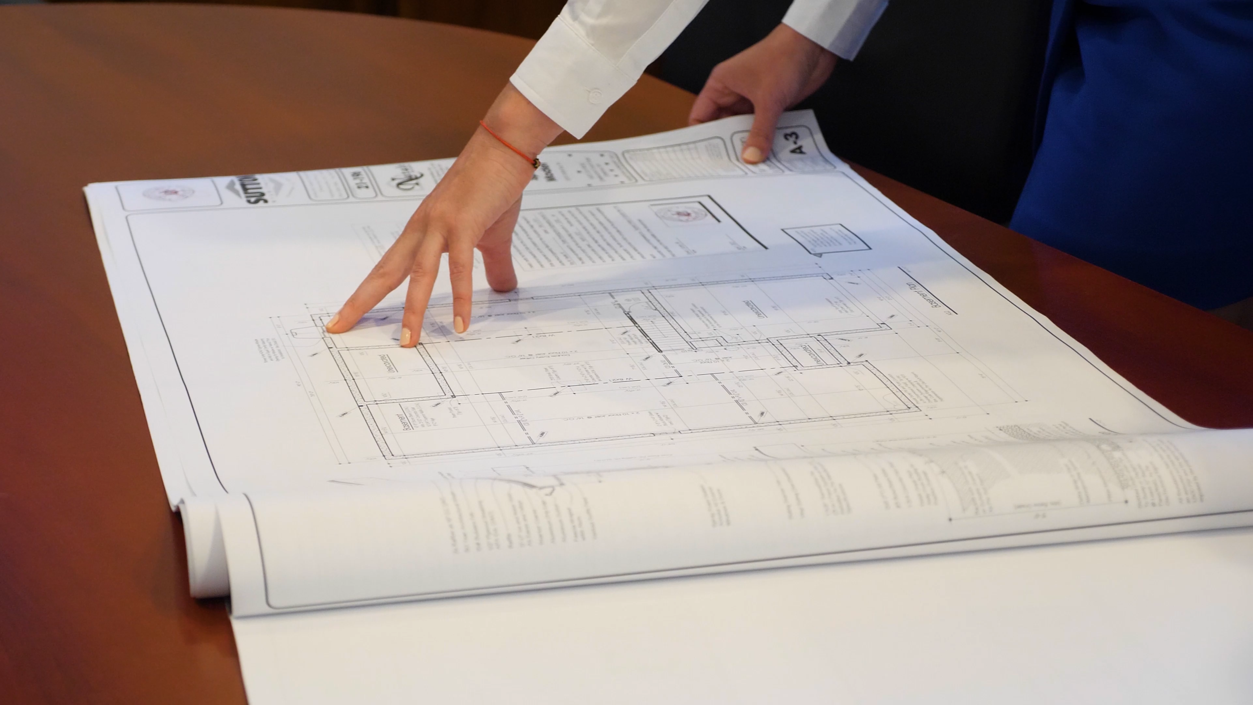 A person pointing at architectural blueprints on a large sheet of paper placed on a wooden table.