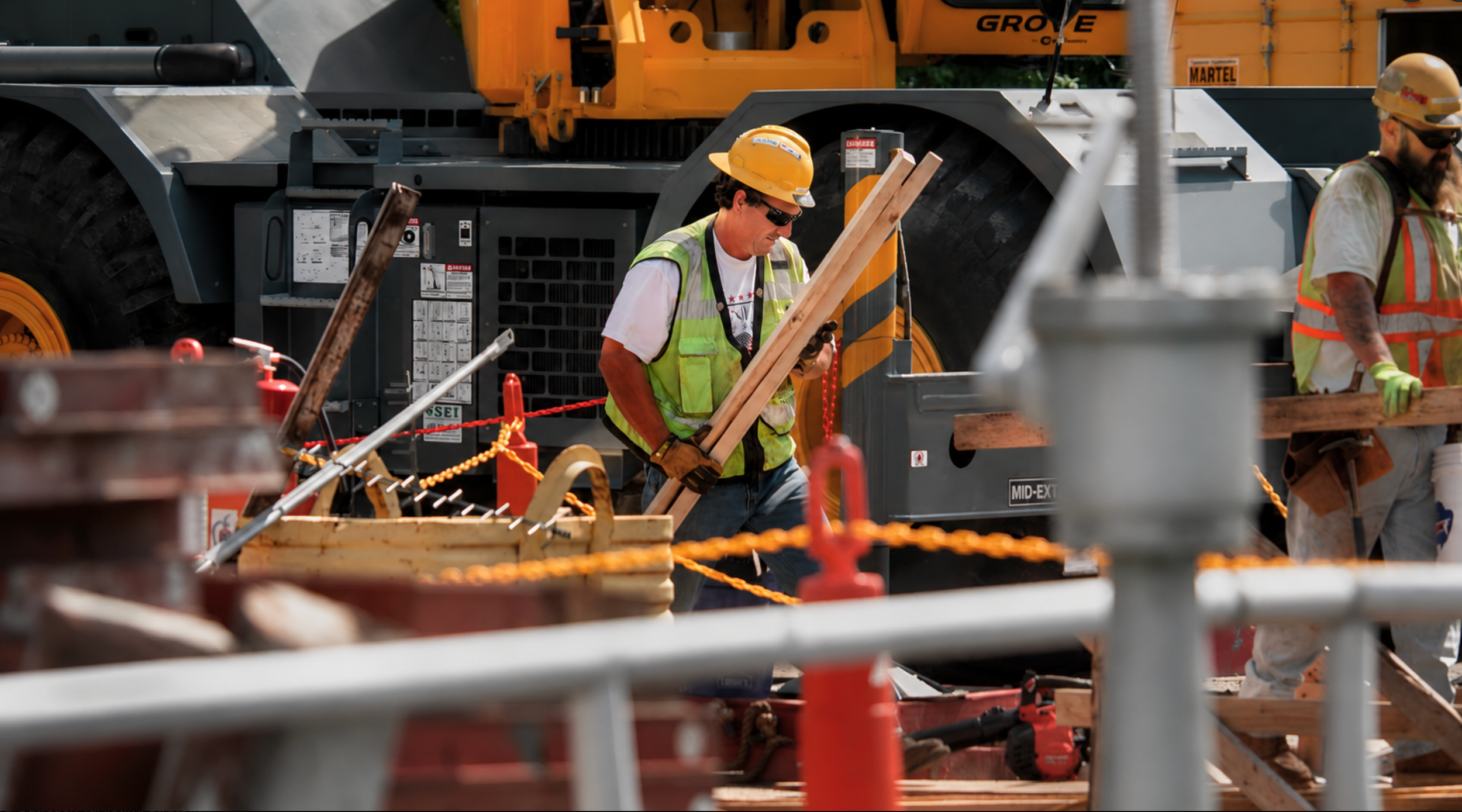 Construction workers wearing yellow hard hats and safety vests working on a site with wooden boards, orange safety ropes, and heavy machinery in the background.