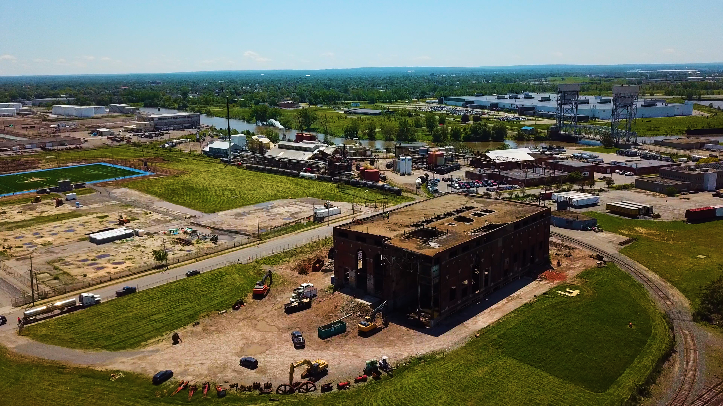 Aerial view of an industrial area with factories, warehouses, and a train bridge crossing a river, with green fields, a sports field, and a mix of vehicles and construction equipment.