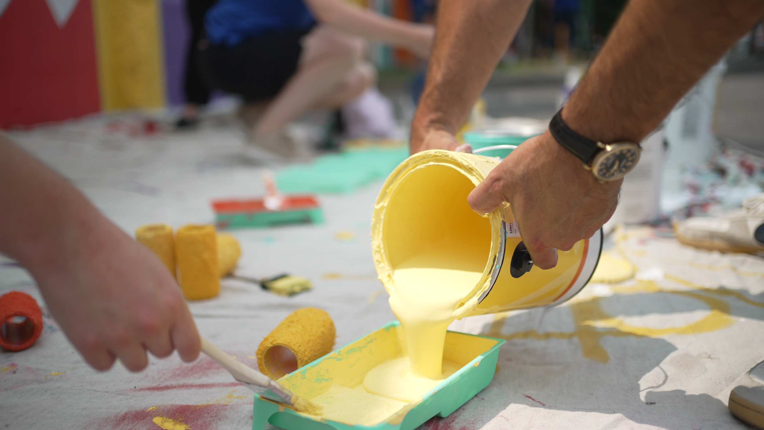 Person pouring yellow paint from a can onto a table with paint rollers nearby, at an outdoor painting activity.