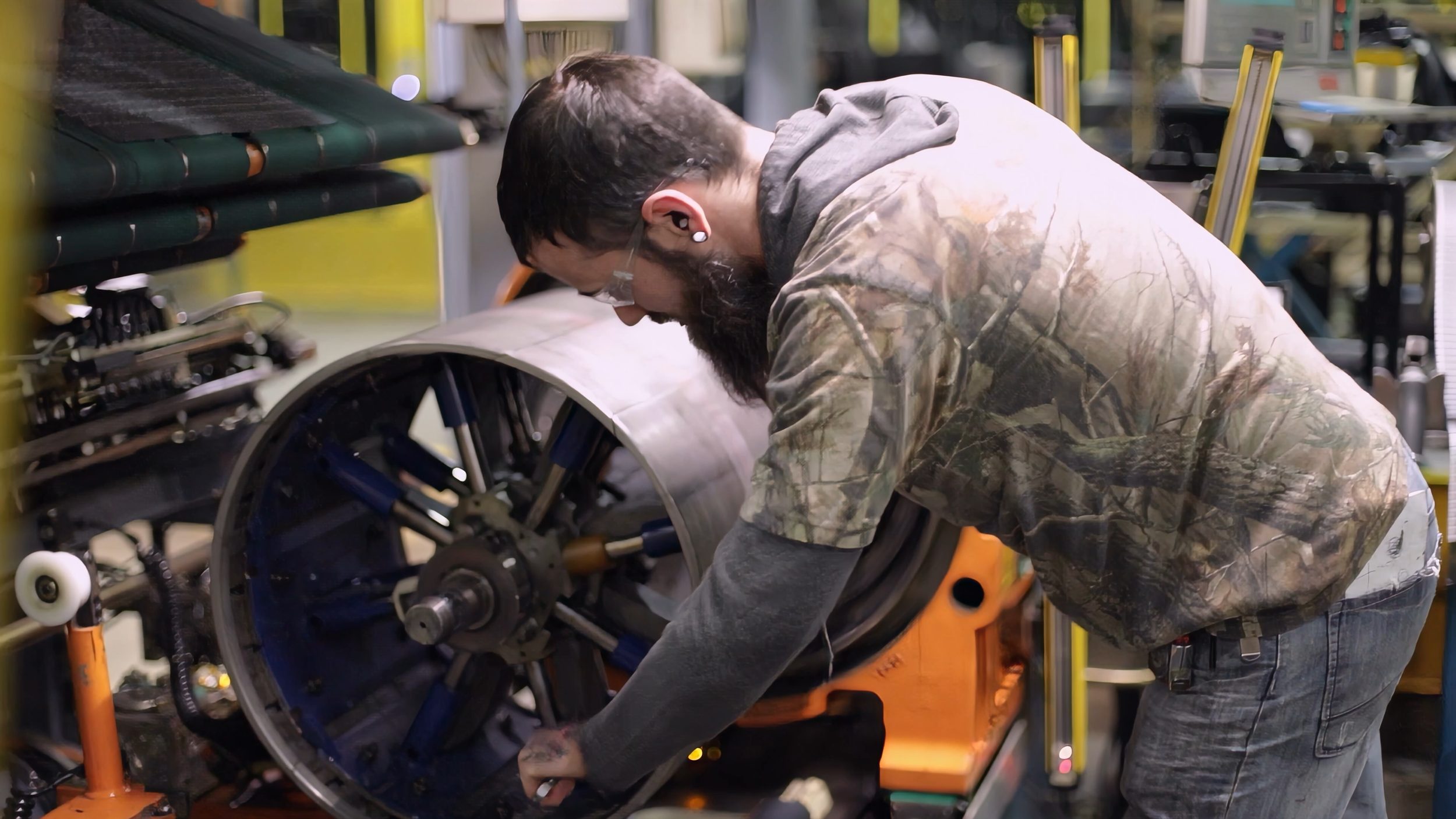 A man working on a large machinery part in a factory or workshop, wearing camouflage jacket and earmuffs.