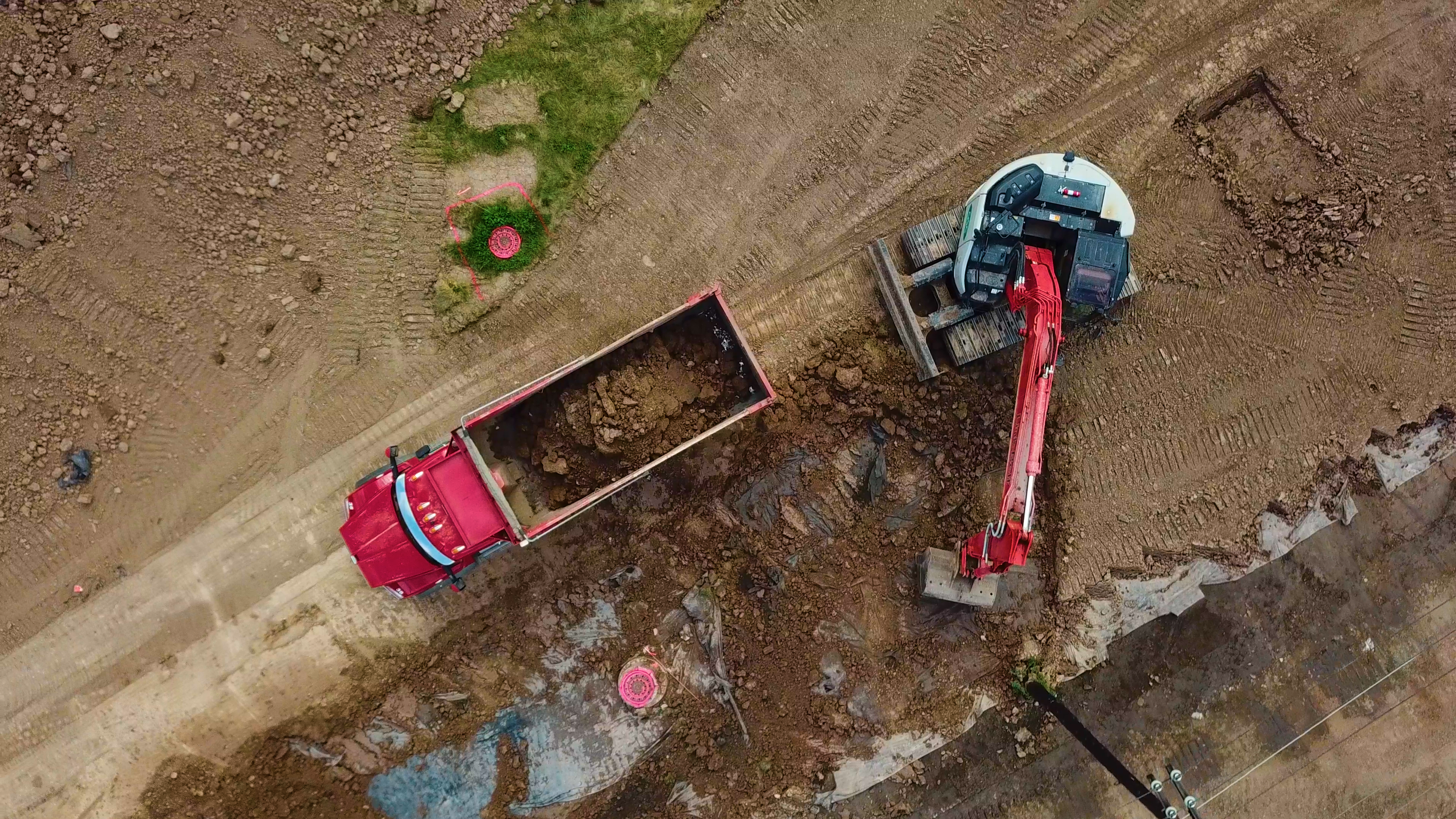 An aerial view of construction equipment including a red dump truck, excavator, and construction site with dirt and ground work.