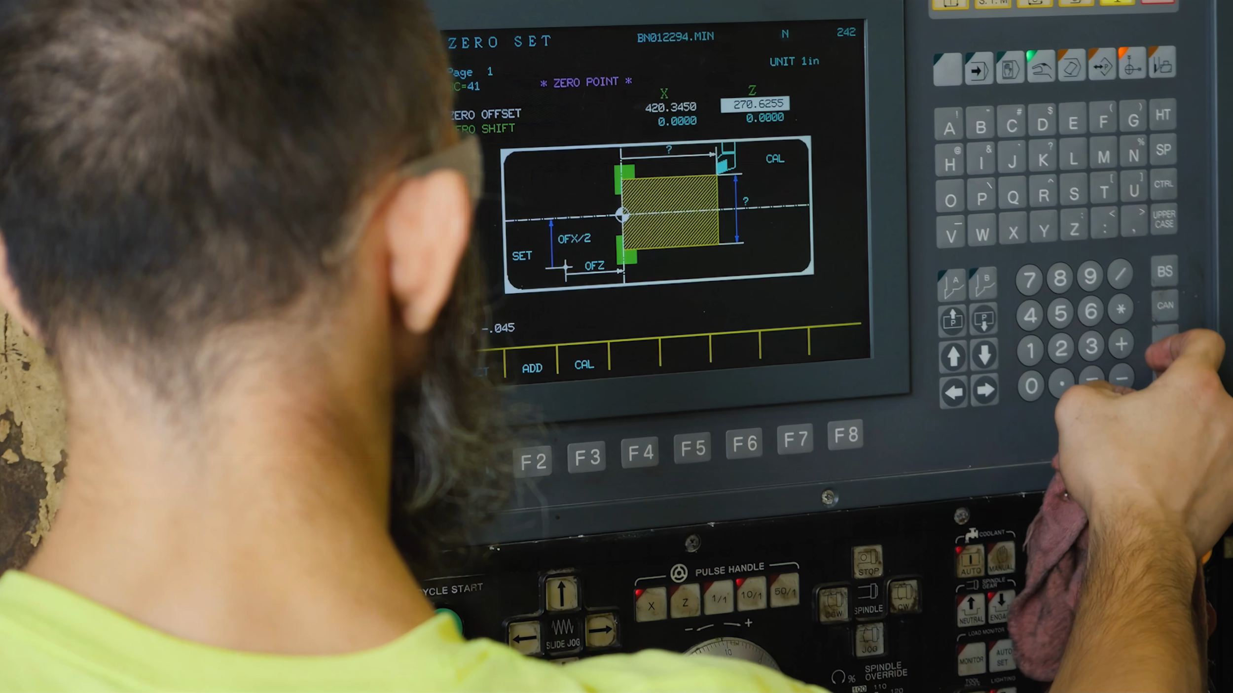 A person with glasses watches a CNC machine screen displaying technical drawings and measurements.