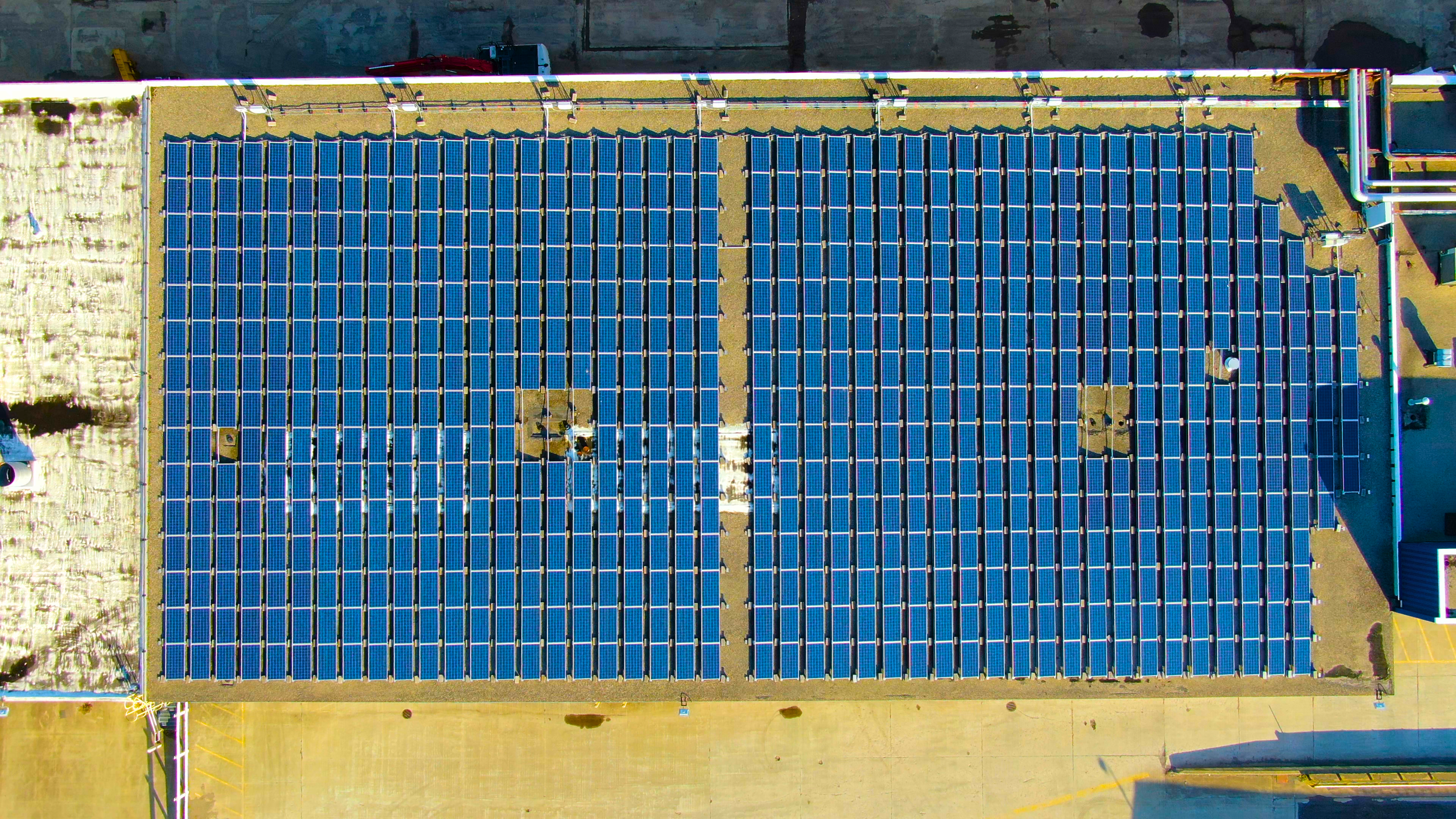A rooftop with multiple blue solar panels arranged in rows, reflecting sunlight, with a section of a parking lot and some buildings visible at the edges.