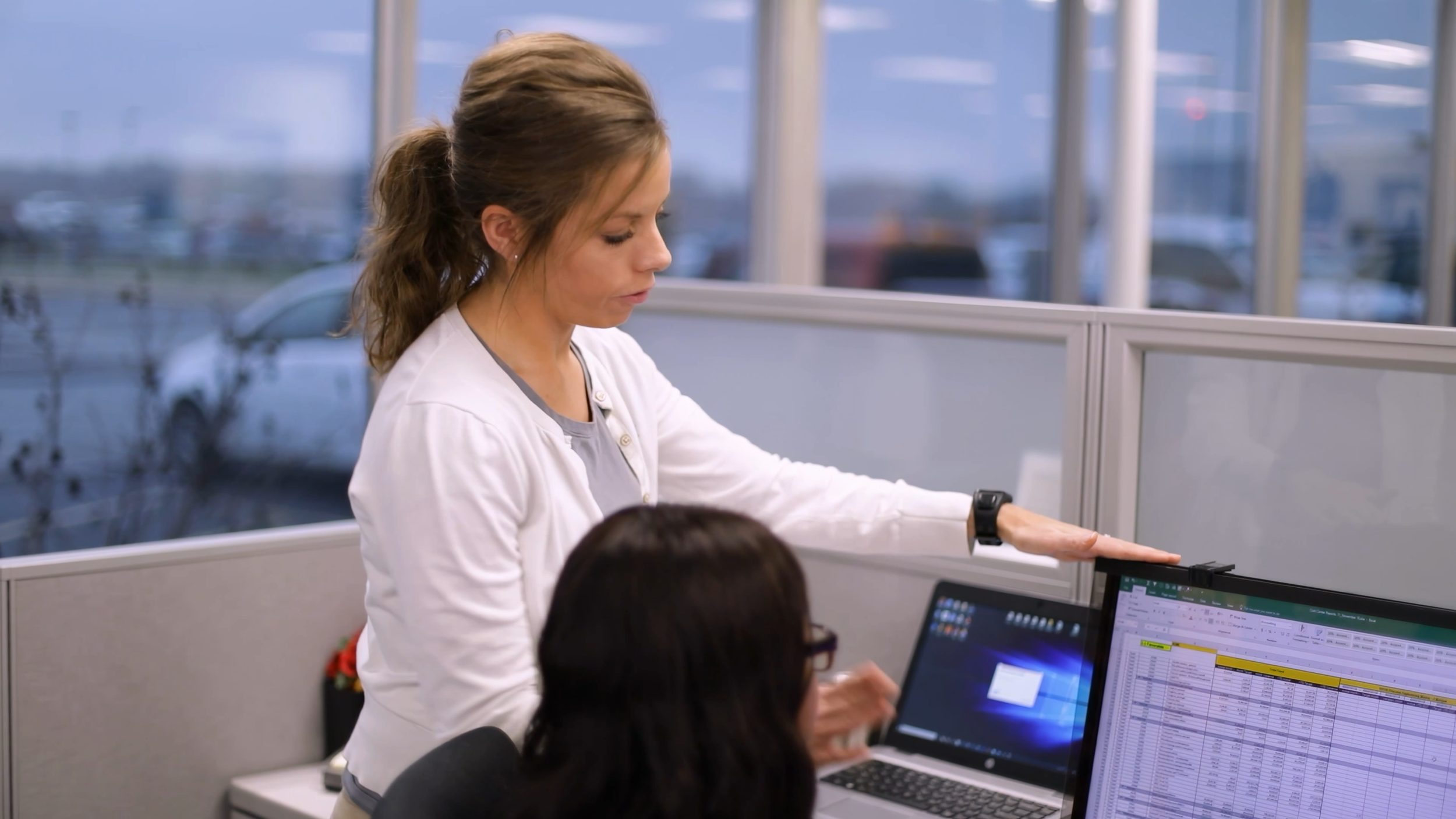 A woman with brown hair tied in a ponytail, wearing a white blouse, is standing at an office cubicle, pointing to a computer screen with spreadsheet data on it. There are other people working at desks nearby, and large windows in the background showing an outdoor parking lot.