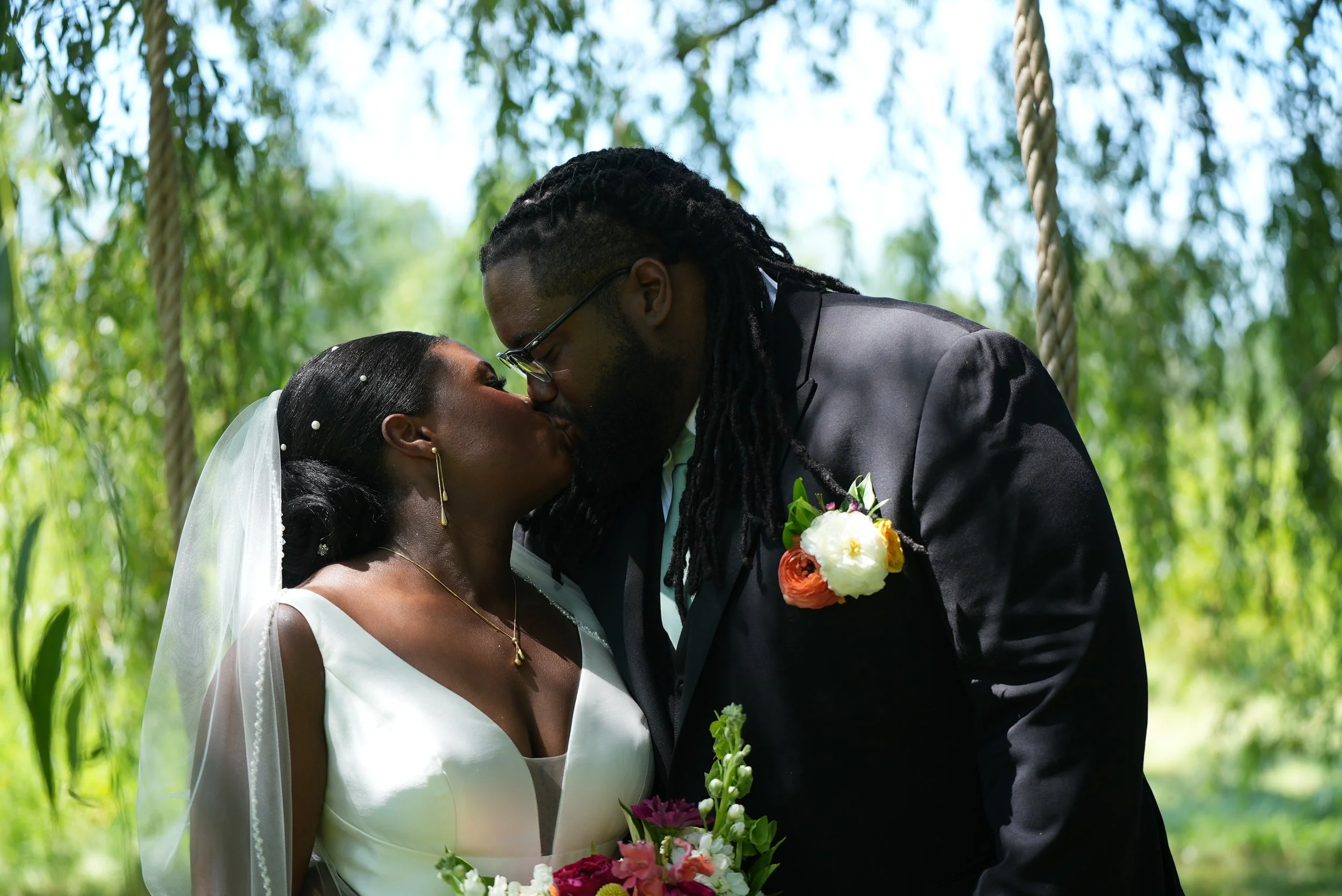A bride and groom kiss during their wedding ceremony outdoors among green trees, with the bride holding a bouquet of flowers.
