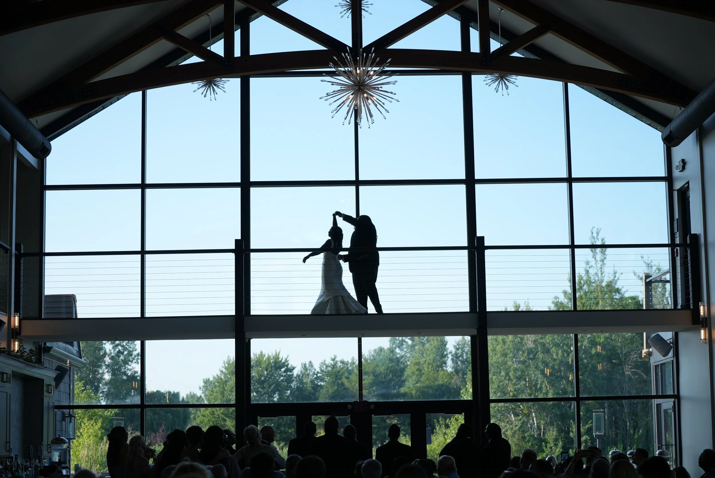 Silhouette of a bride and groom dancing on a balcony inside a building with large glass windows and ceiling lights, with an audience seated below watching.