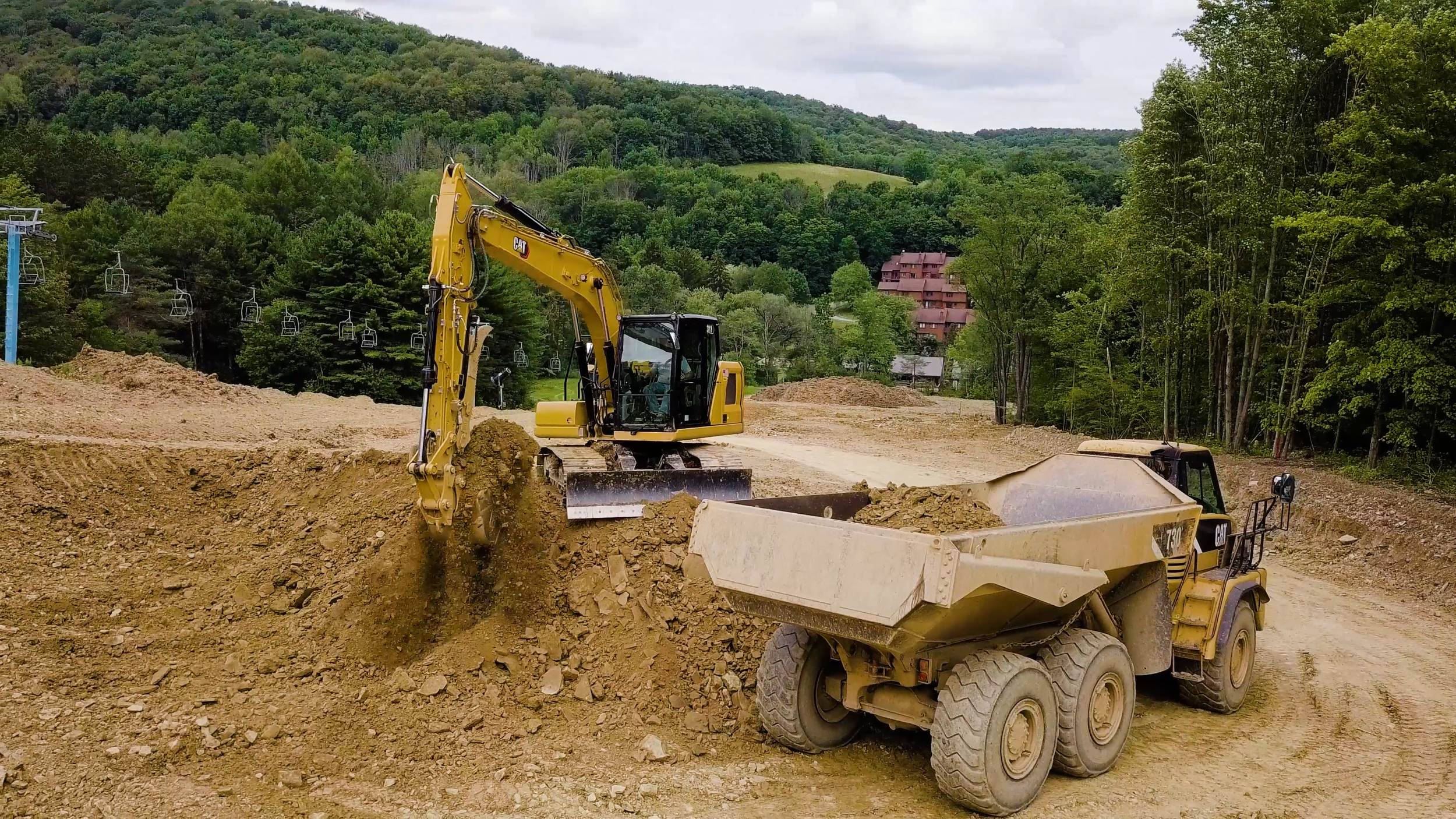 A yellow excavator is digging and moving dirt on a construction site surrounded by trees and rolling hills.