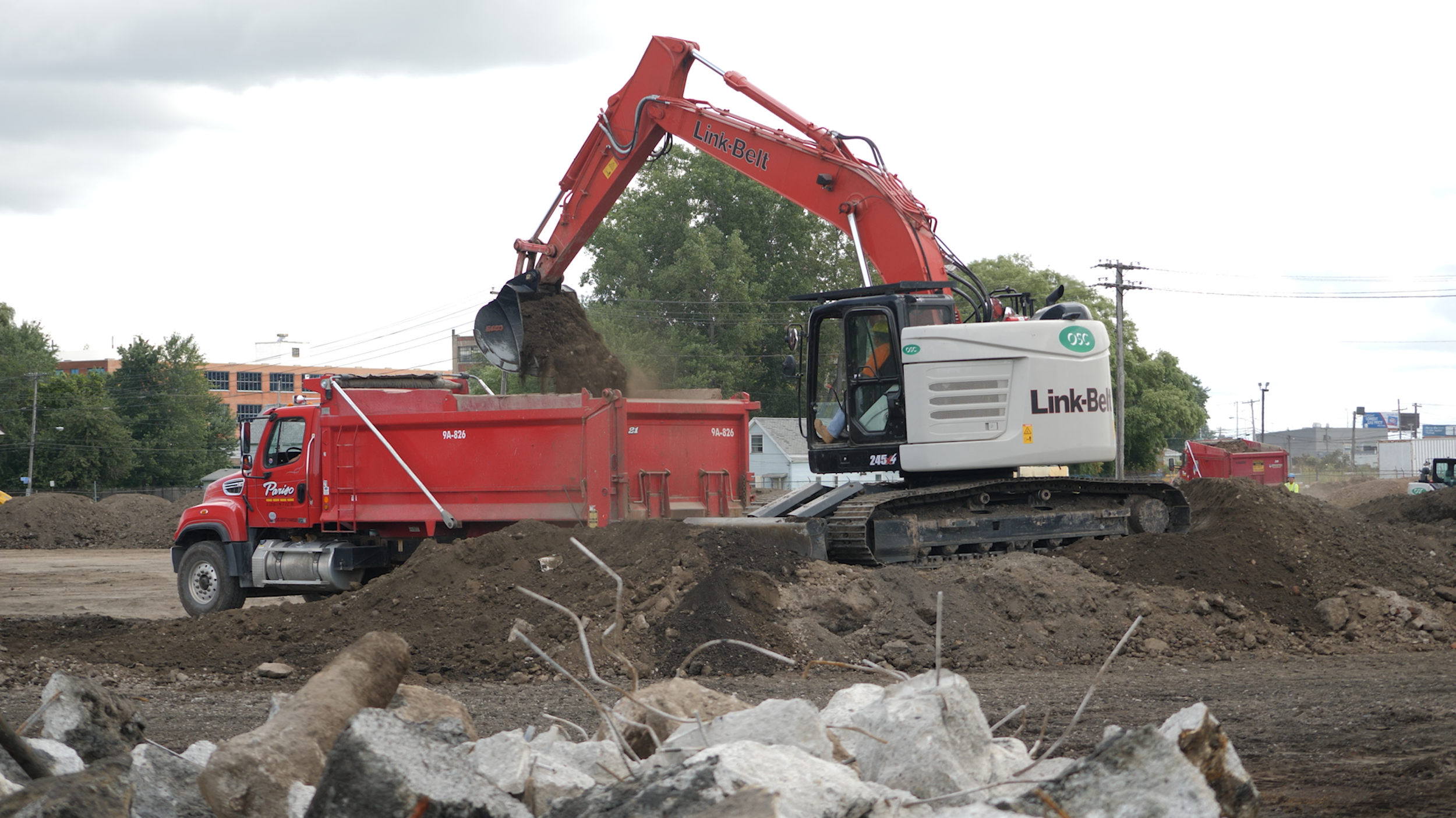 An orange and white excavator digging dirt at a construction site with a red dump truck next to it and buildings in the background.