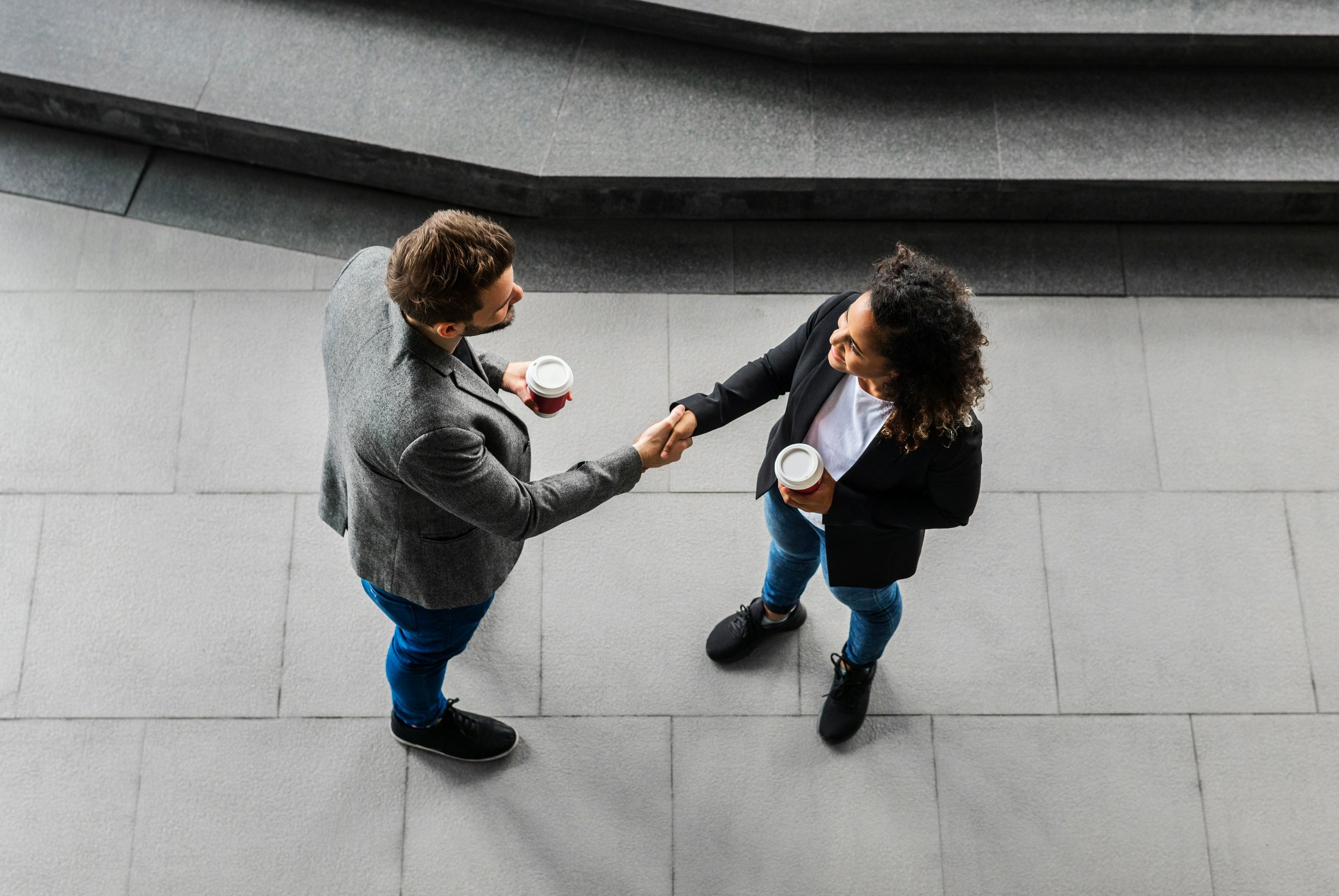 Top-down view of a man and a woman shaking hands while holding coffee cups, standing on a tiled floor.