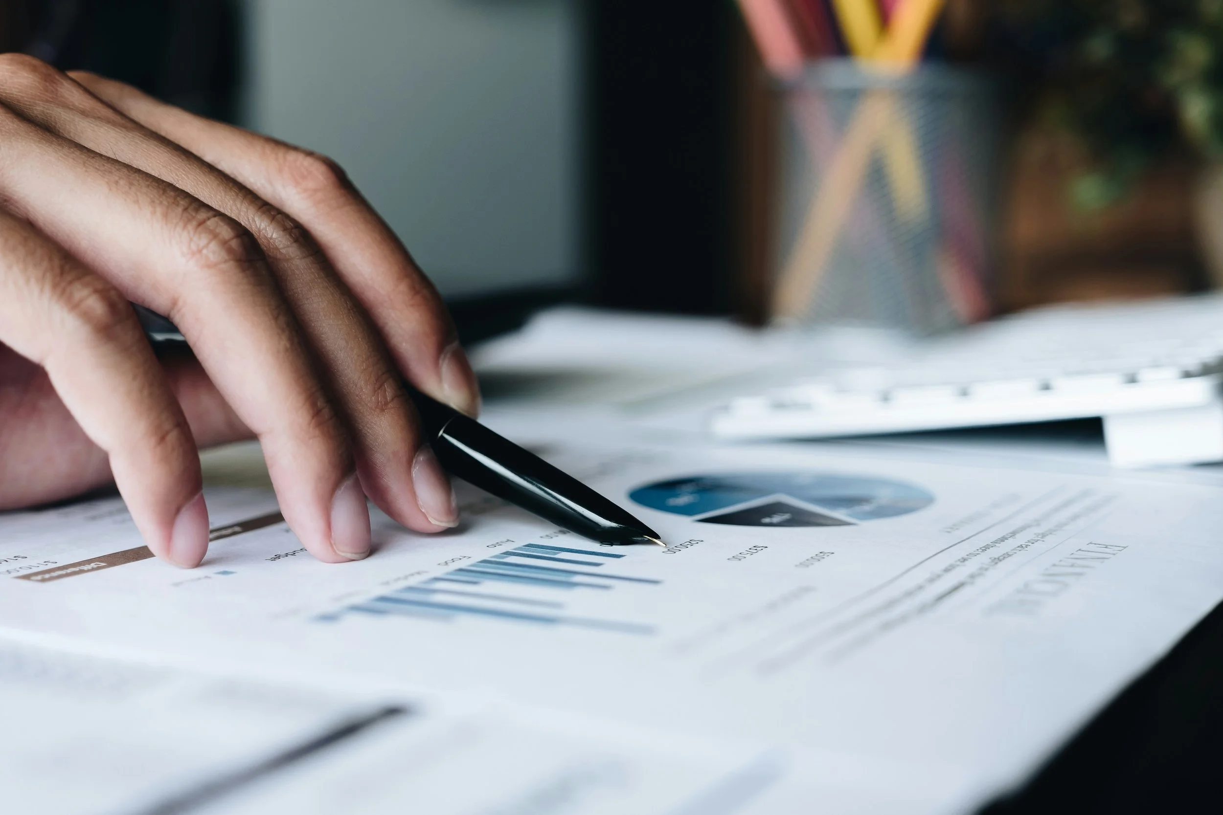 Close-up of a person's hand writing on financial documents with a black pen on a desk, with charts and graphs visible on the paper, and a blurred background of stationery in a striped container.