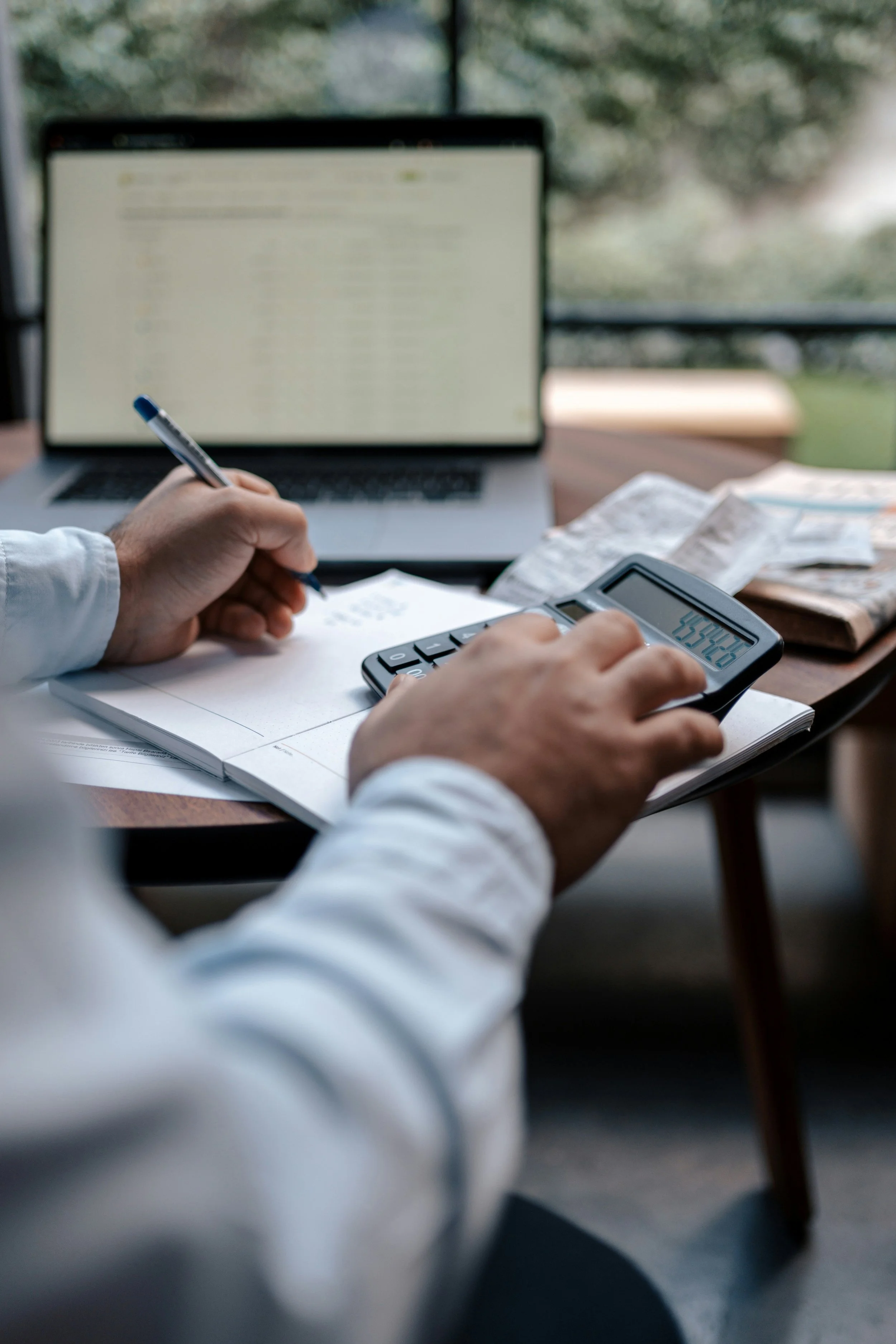 Person in white shirt using a calculator, with a pen in hand and papers on desk, in front of a laptop with blurred screen, on a desk with newspapers, near a window with trees outside.
