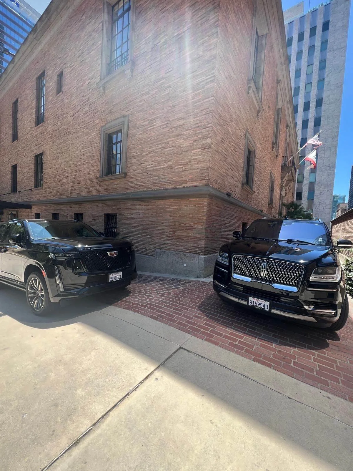 Black Cadillac and black Lincoln SUVs parked on a brick-paved area beside a brick building with tall windows during the daytime.