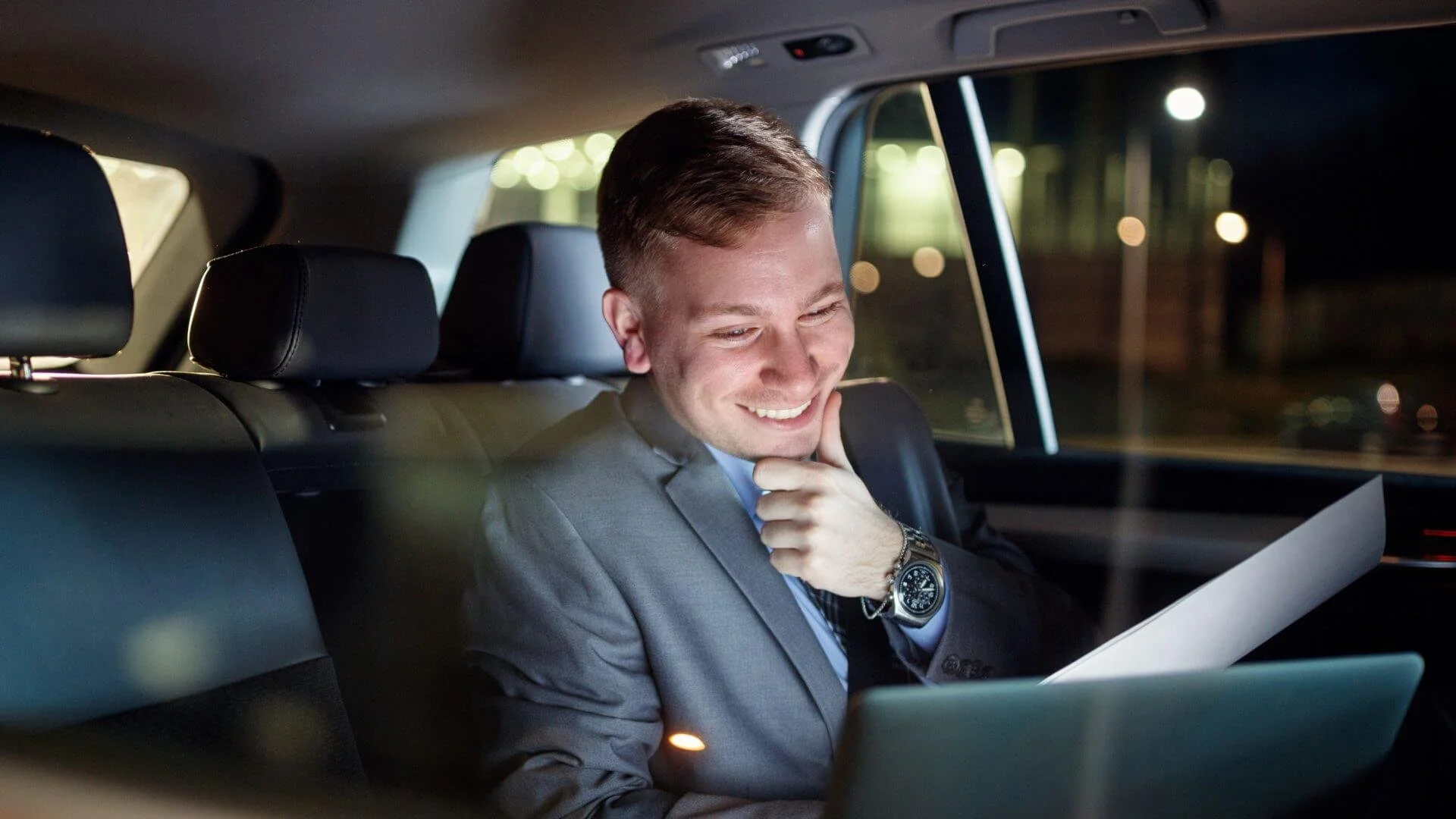 A man in a suit sitting in the back seat of a car, smiling and looking at a tablet. It is nighttime outside the car.