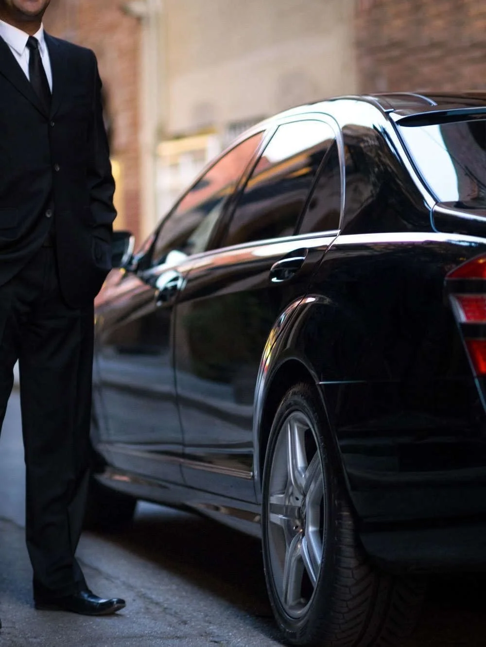 A man in a black suit standing next to a black luxury car with tinted windows.