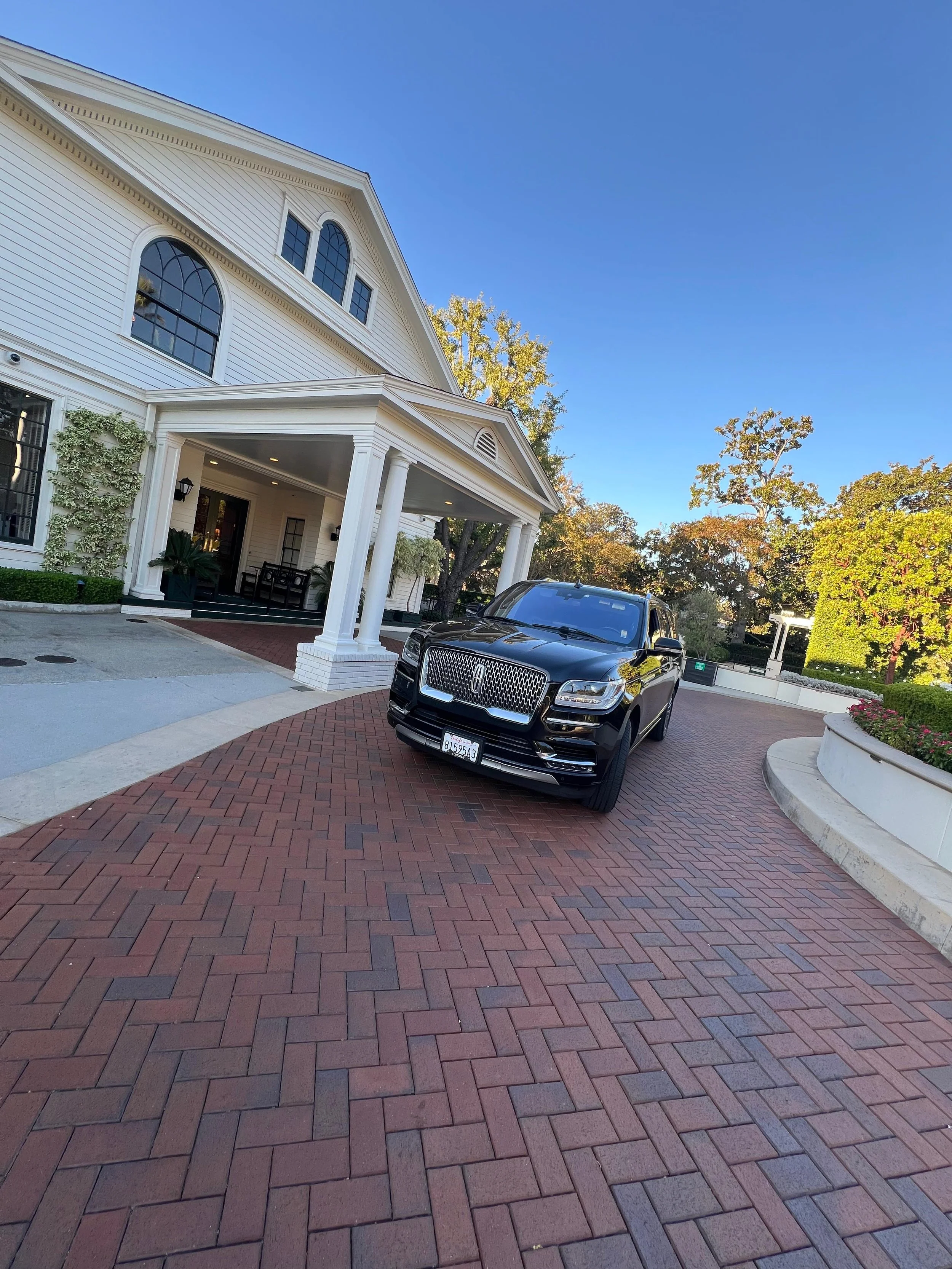 A black luxury SUV parked on a brick driveway in front of a large white house with columns and arched windows, surrounded by trees and greenery.