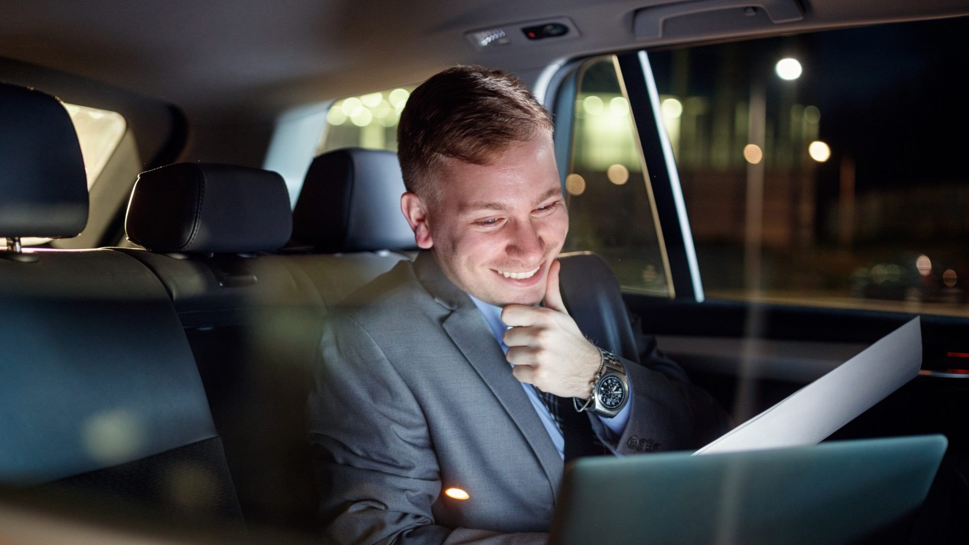 A man in a suit sitting in the back seat of a car, smiling and looking at a tablet. It is nighttime outside the car.