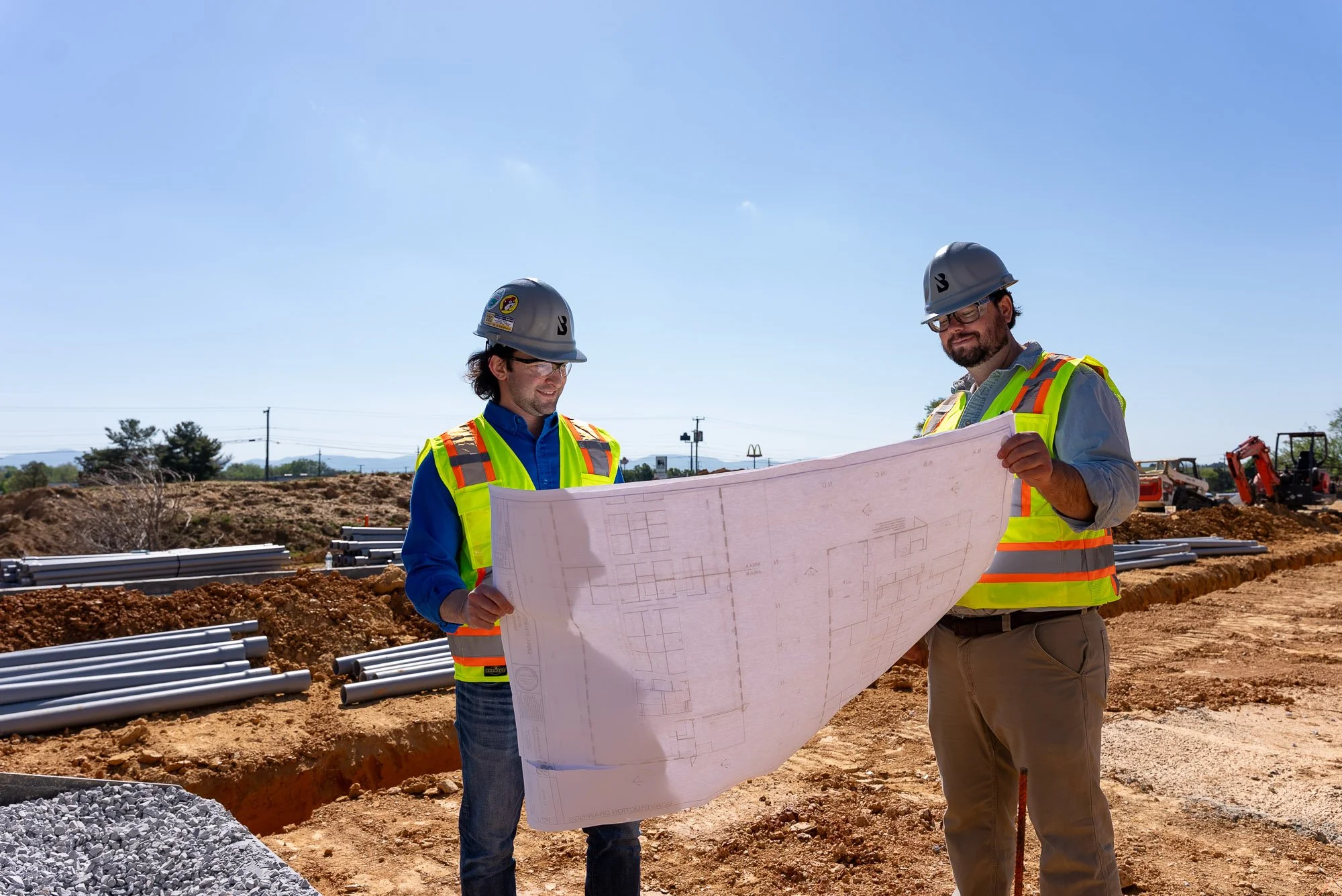 Two construction workers in safety vests and helmets looking at a blueprint on a construction site with pipes and machinery in the background.