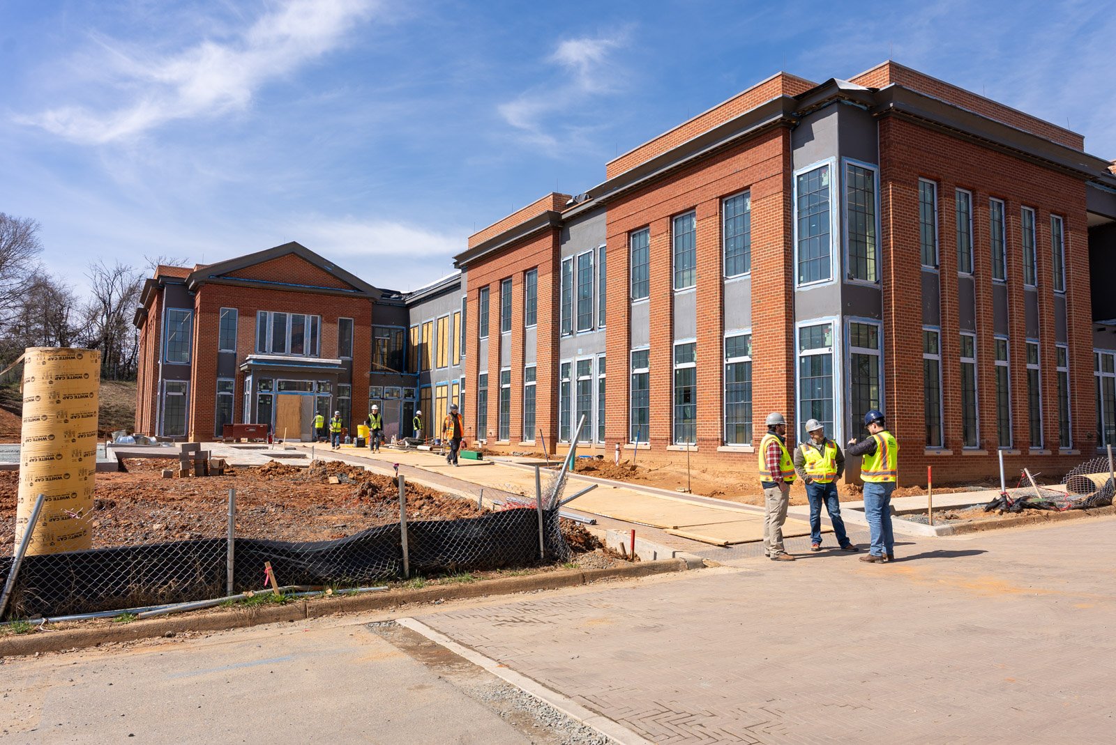 Construction workers in safety vests and helmets working on a large brick building under construction, with scattered construction materials and equipment around.