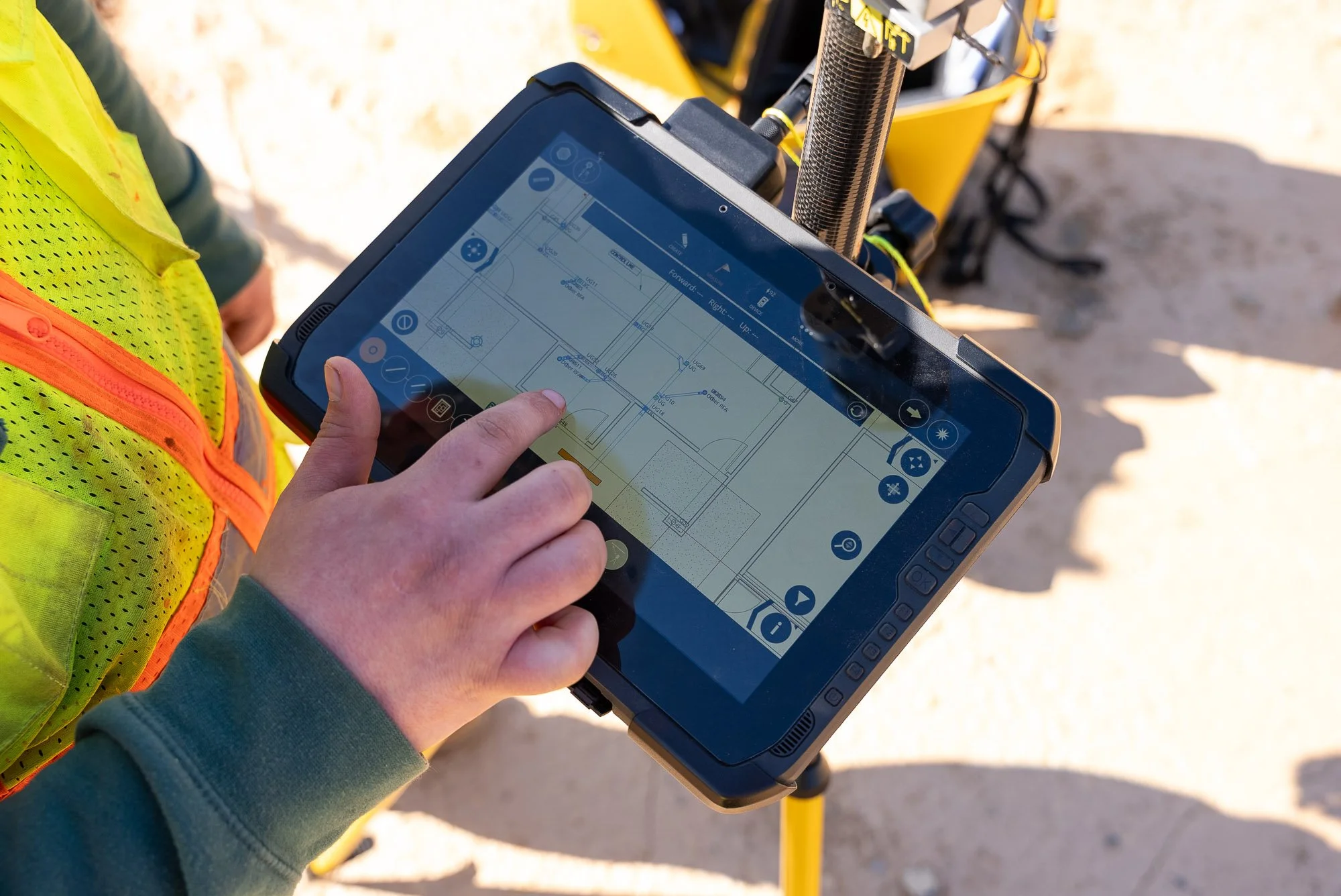 A person holding a tablet displaying a technical drawing or blueprint, with a construction or engineering site in the background, including a yellow piece of machinery and a person wearing a high-visibility safety vest.