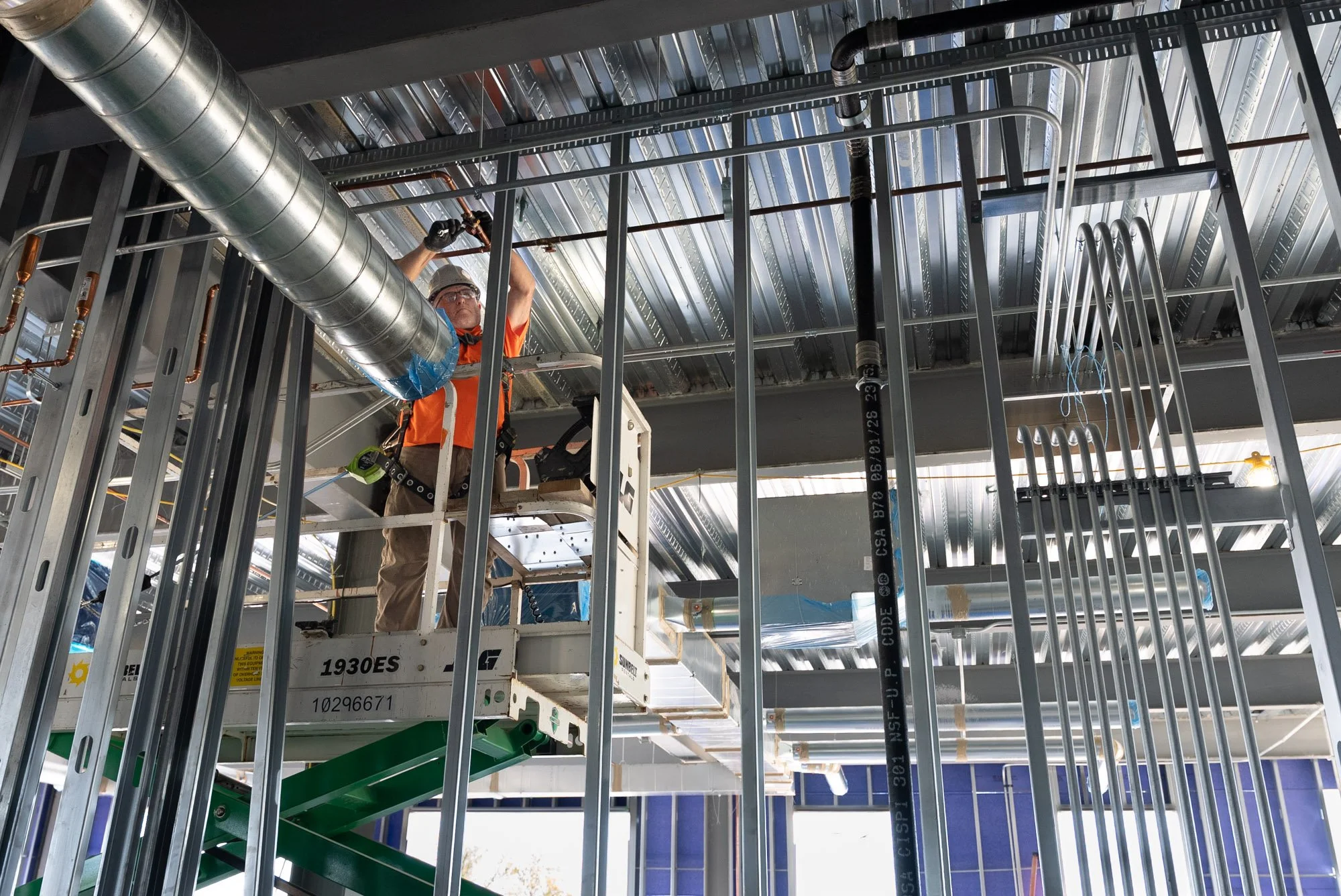 A mechanical contractor technician on a scissor lift connecting copper piping beside spiral ductwork and cast iron plumbing at the Daikin Applied office building in Verona, VA.