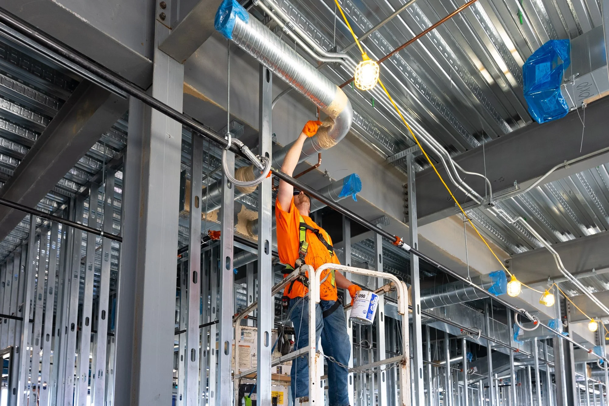 A Blauch Brothers technician on a scissor lift sealing an overhead duct and copper pipe connection during commercial HVAC and piping installation at the Daikin Applied office building in Verona, VA.