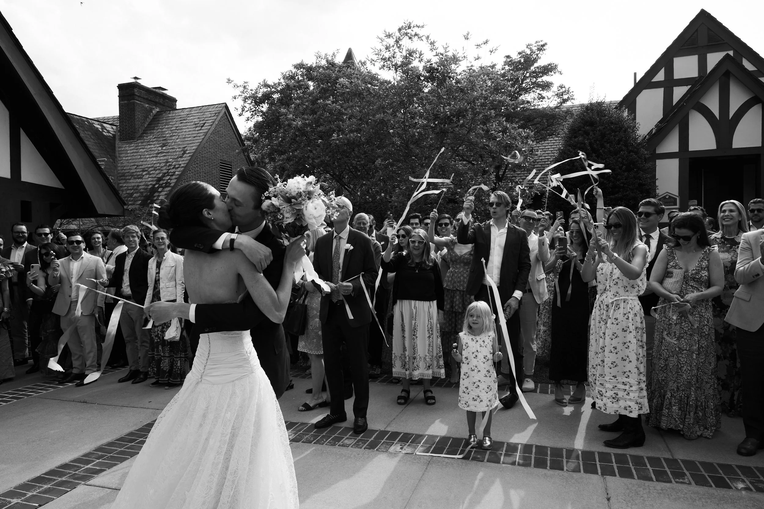 A newlywed couple sharing a kiss, the bride holding a bouquet, surrounded by cheering guests at an outdoor wedding celebration.