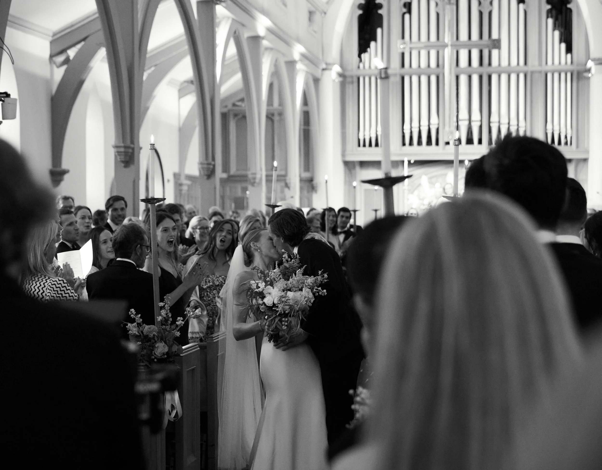 A black and white photo of a wedding ceremony inside a church with a bride and groom kissing, surrounded by guests, lit candles, and a large pipe organ in the background.