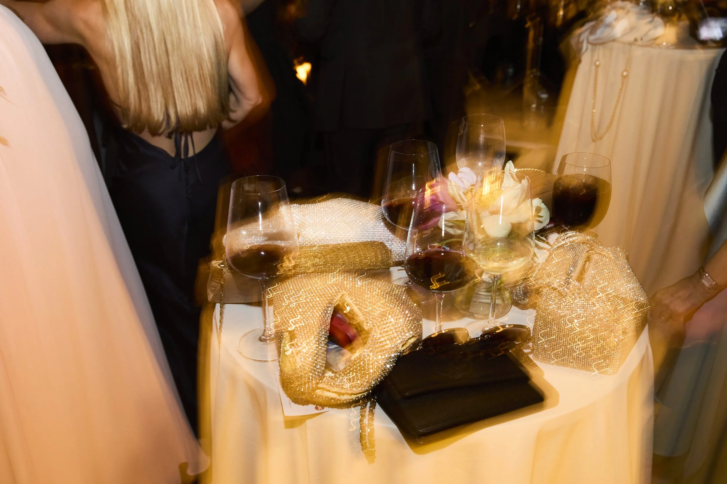 A table set at an event with three glasses of red wine, decorated with golden mesh gift boxes, a string of white flowers, and a black wallet, with guests standing around.