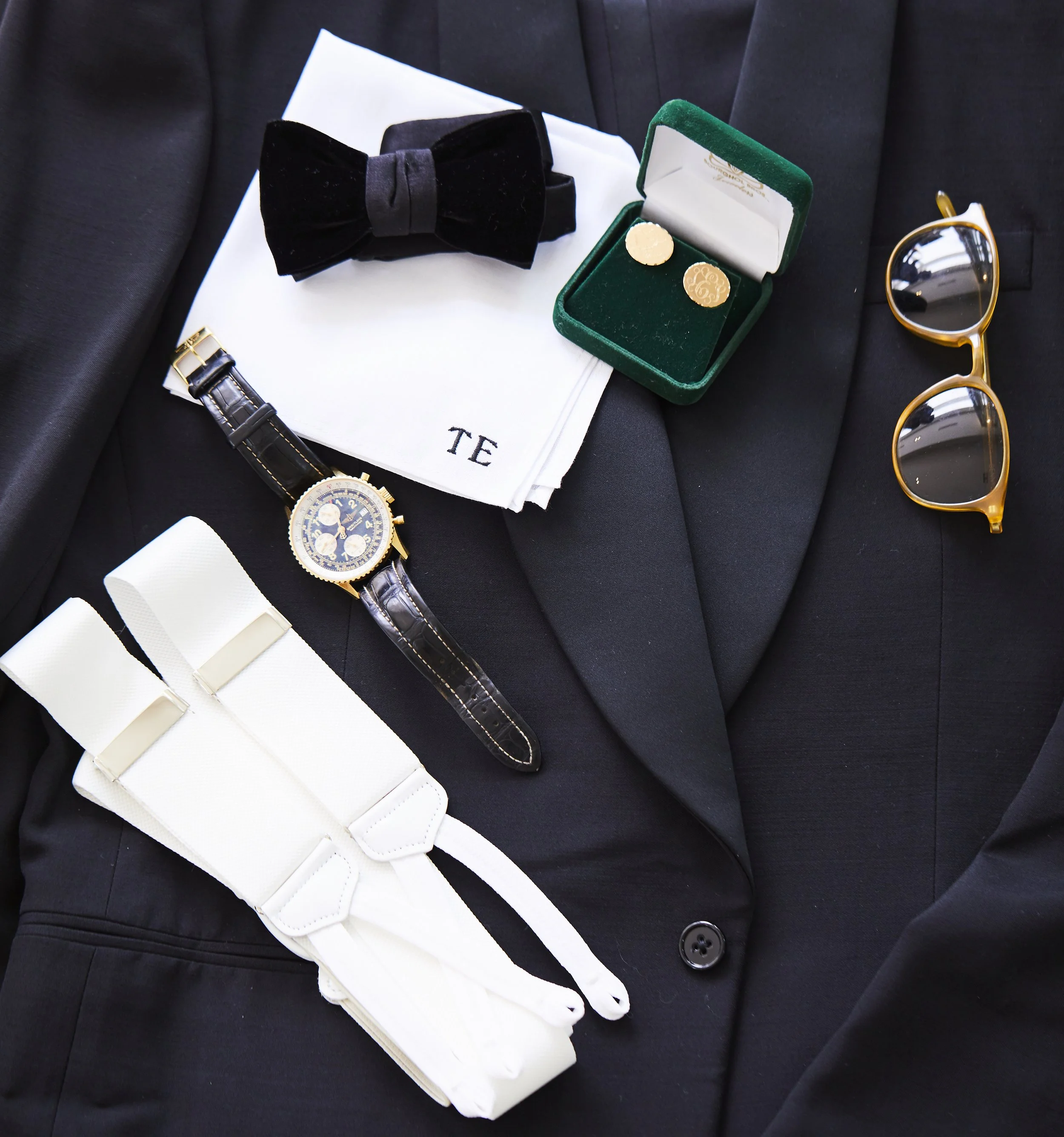 Elegant men's formal accessories arranged on a black tuxedo jacket, including a black bow tie, a pair of gold sunglasses, a wristwatch with a black leather strap, gold cufflinks, a white tuxedo shirt with French cuffs, and a white pocket square.