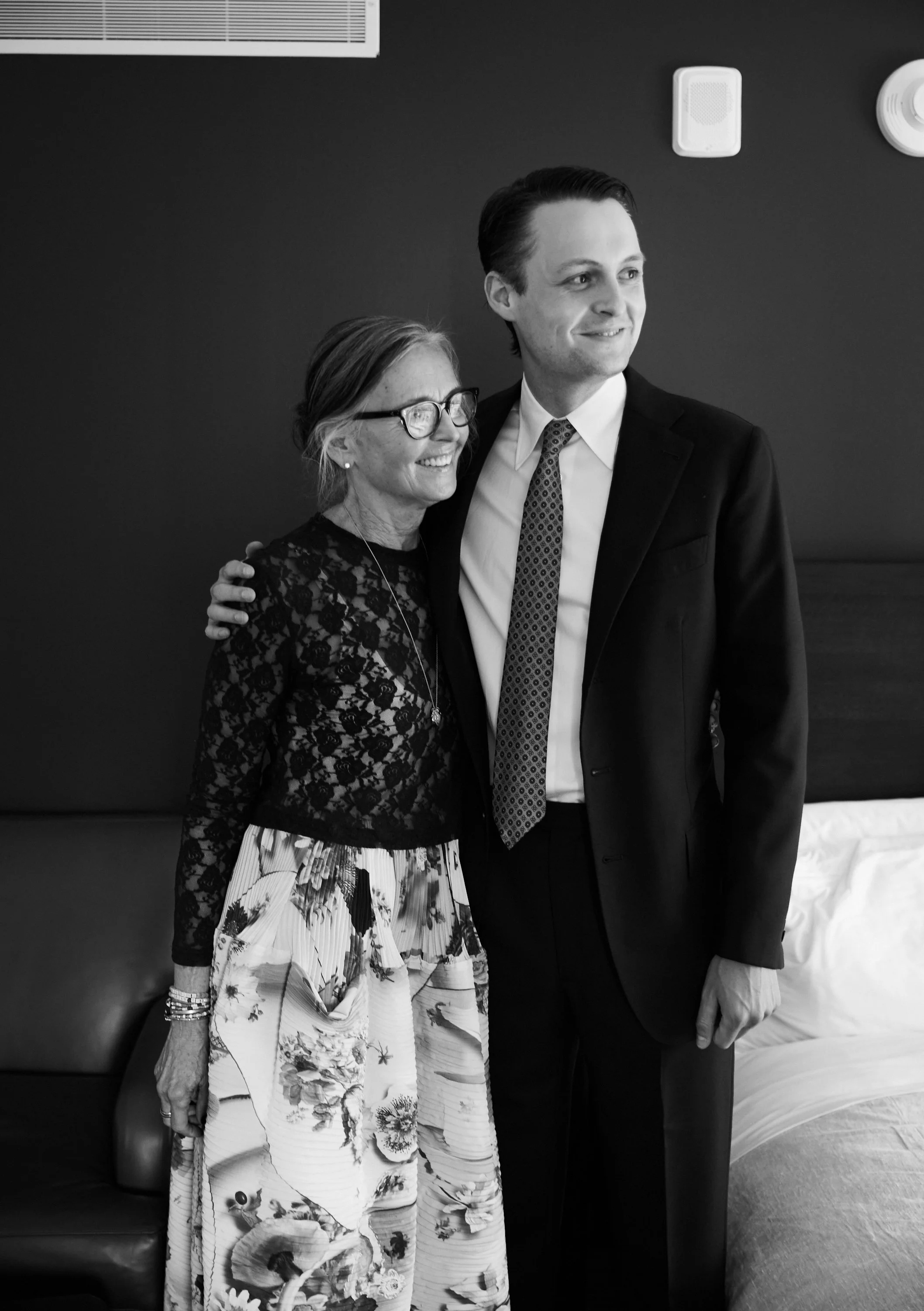 A woman and a man are posing together in a black-and-white photograph. The woman, wearing glasses and a floral skirt with a lace top, is smiling. The man, dressed in a suit and tie, has his arm around her shoulder and is also smiling.
