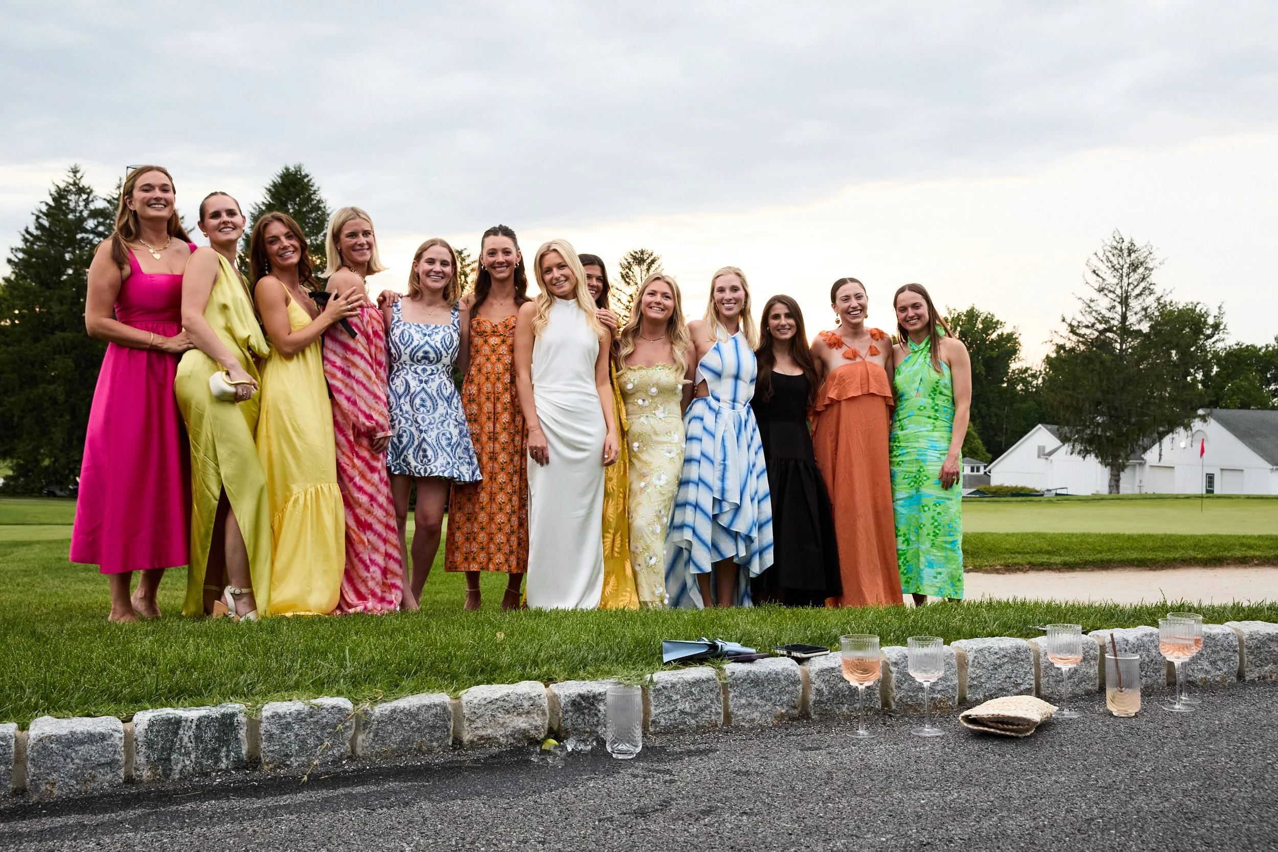 Group of young women dressed in colorful dresses standing together outdoors on a green lawn, smiling for a photo during an event, with glasses of rosé and a purse on the ground in front of them.