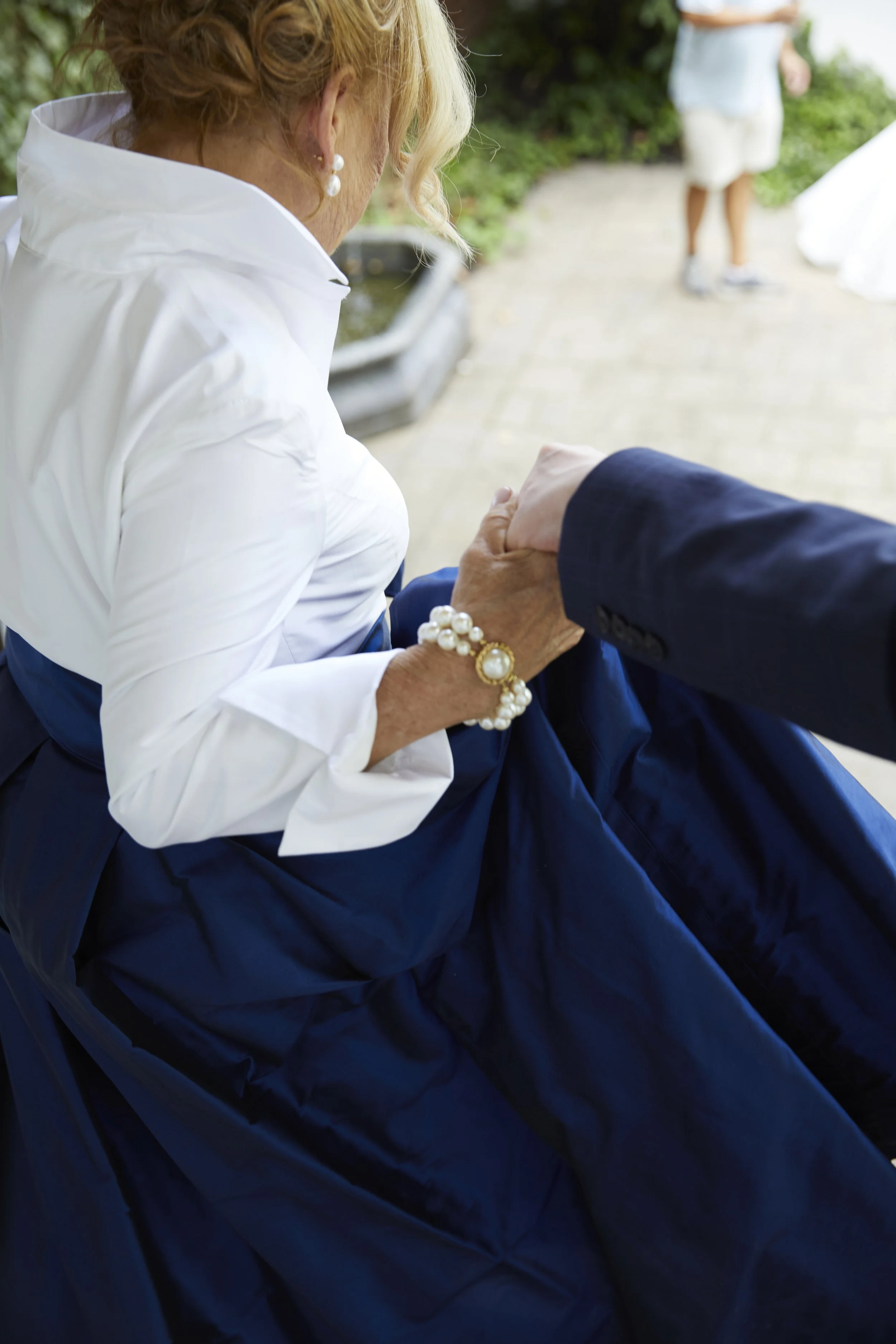 An elderly woman dressed in a white blouse and a long navy blue skirt is holding hands with a man. She is wearing pearl jewelry including earrings, a bracelet, and a ring. They are outdoors on a paved path with greenery in the background, and a perso
